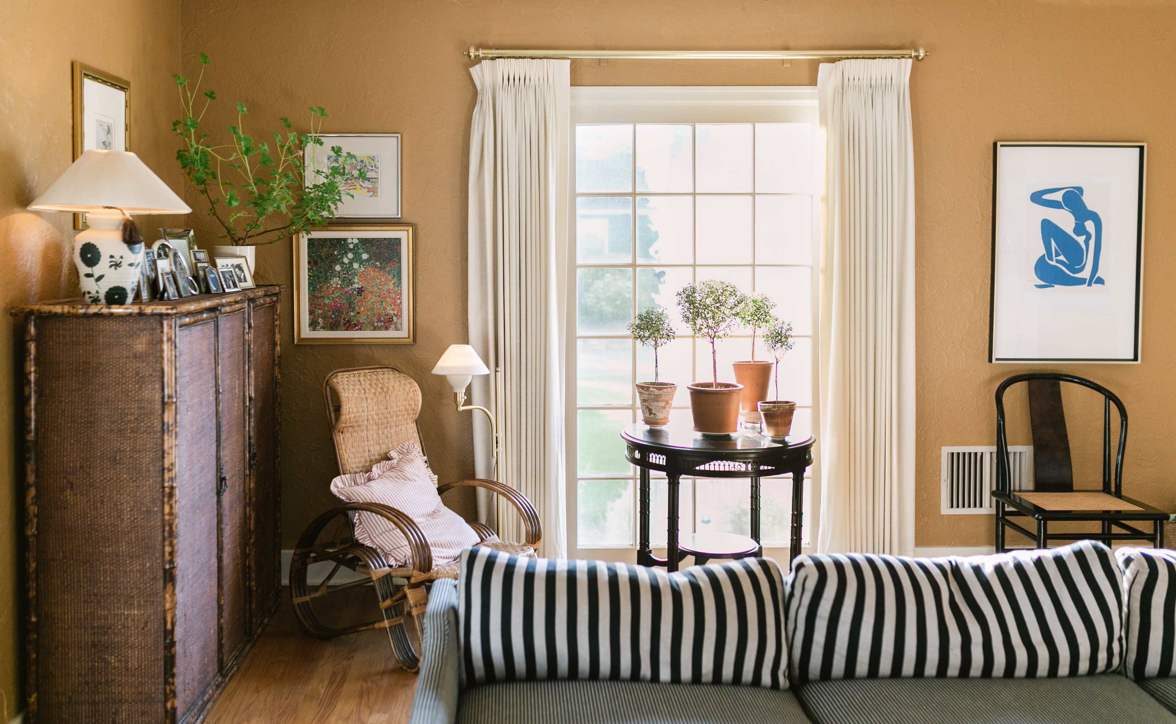 A cozy living room features a striped sofa, a wicker rocking chair, potted plants on a round table, and artworks on the walls.