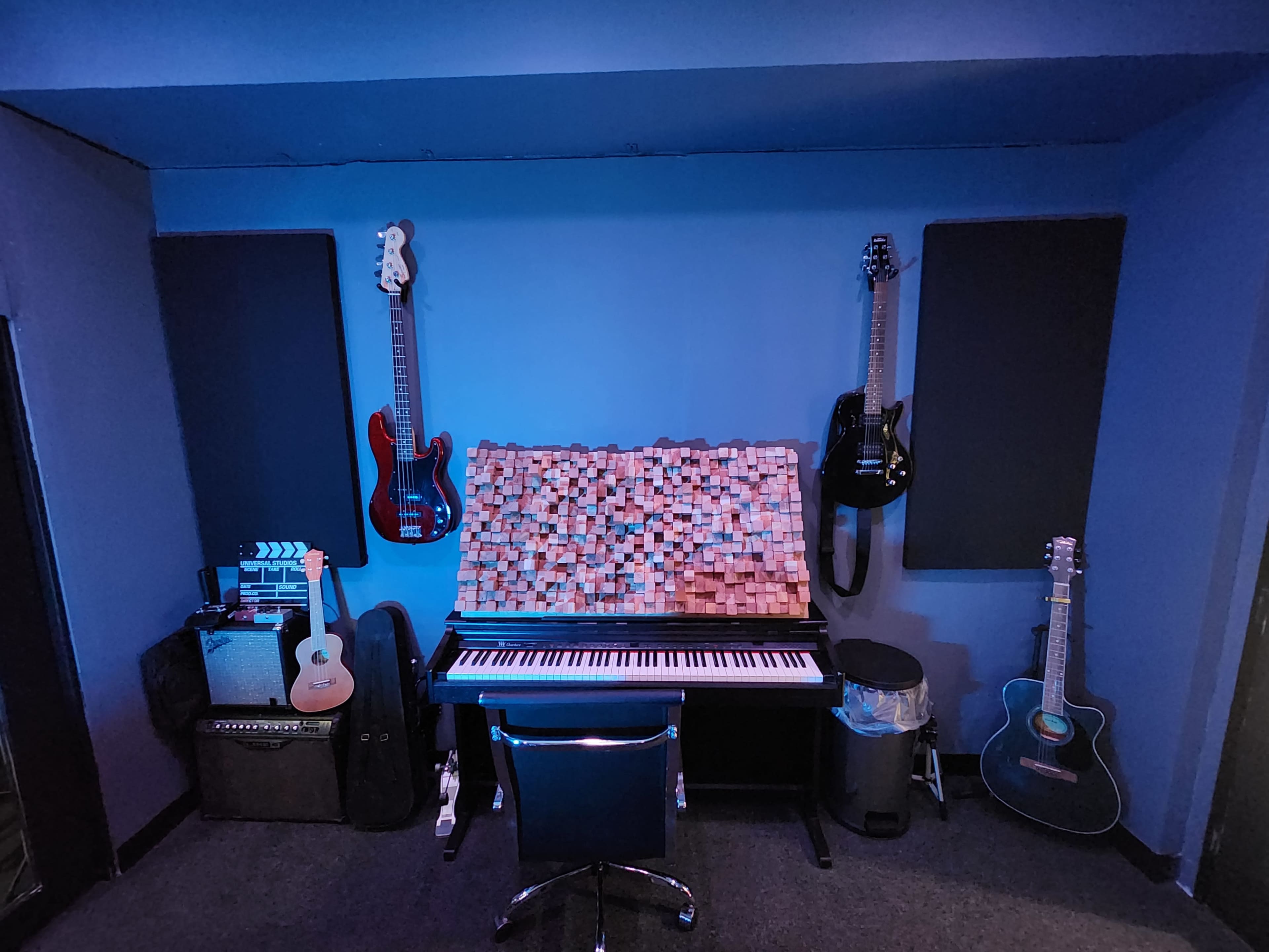 A music studio setup featuring a keyboard at the center, surrounded by various guitars hung on the walls, acoustic panels, and amplifiers.