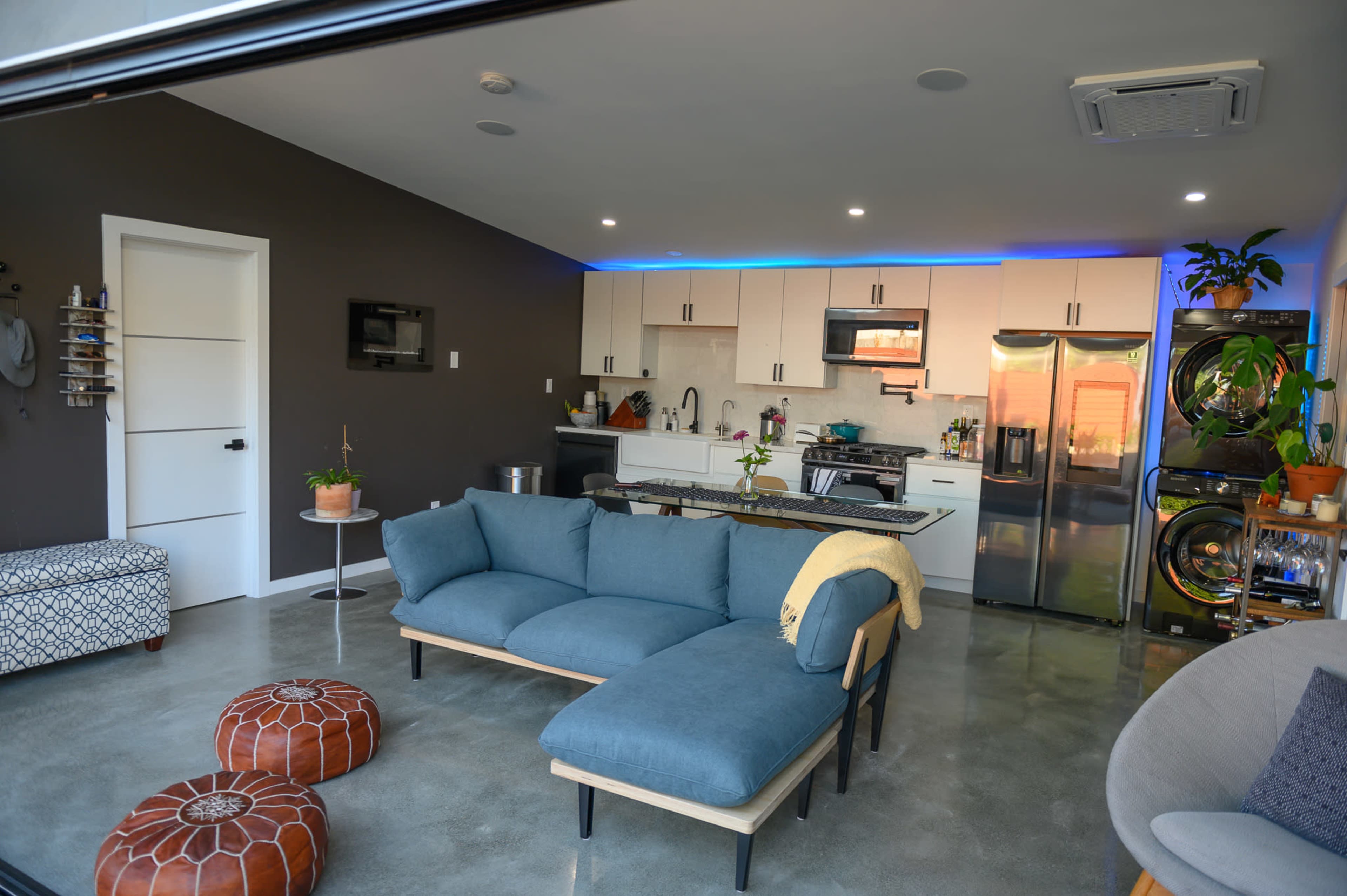 A modern living space features a blue sectional sofa, a kitchen with light-colored cabinets and stainless steel appliances, and decorative plants.