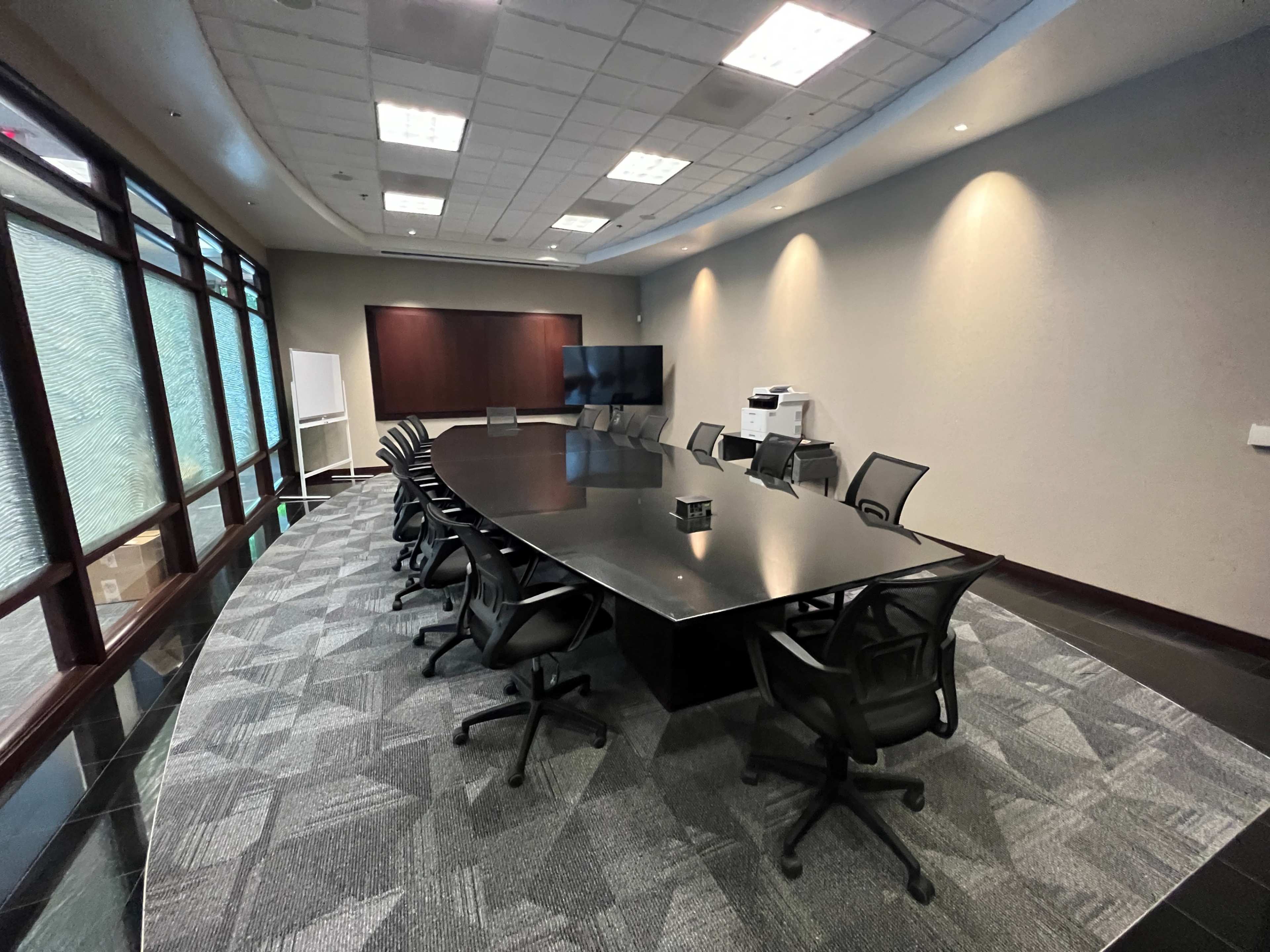 A modern conference room features a large black table surrounded by mesh office chairs, with a television screen and whiteboard against one wall.