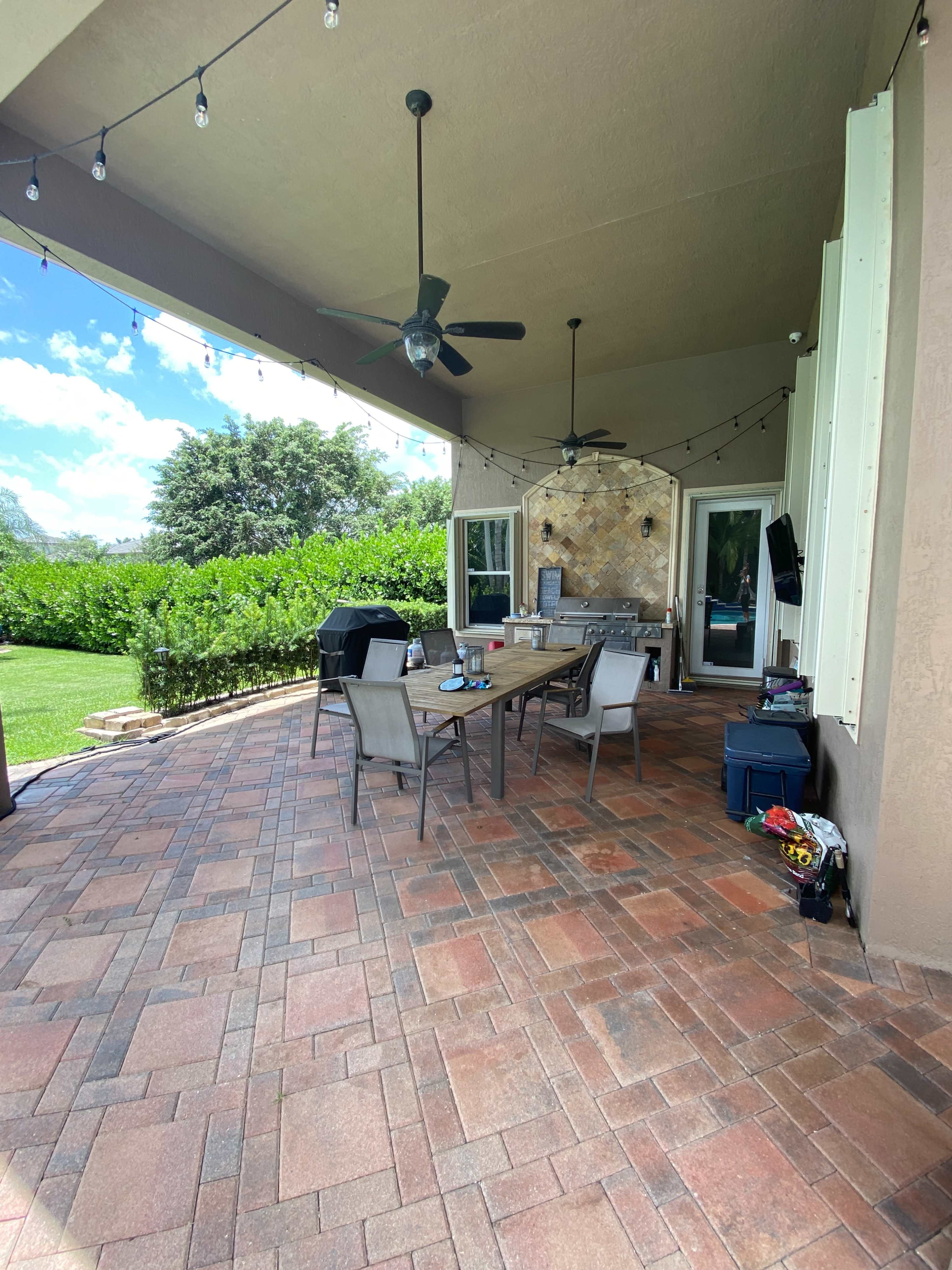 A covered patio features a long table surrounded by chairs, with a grill and outdoor seating area in a landscaped garden.