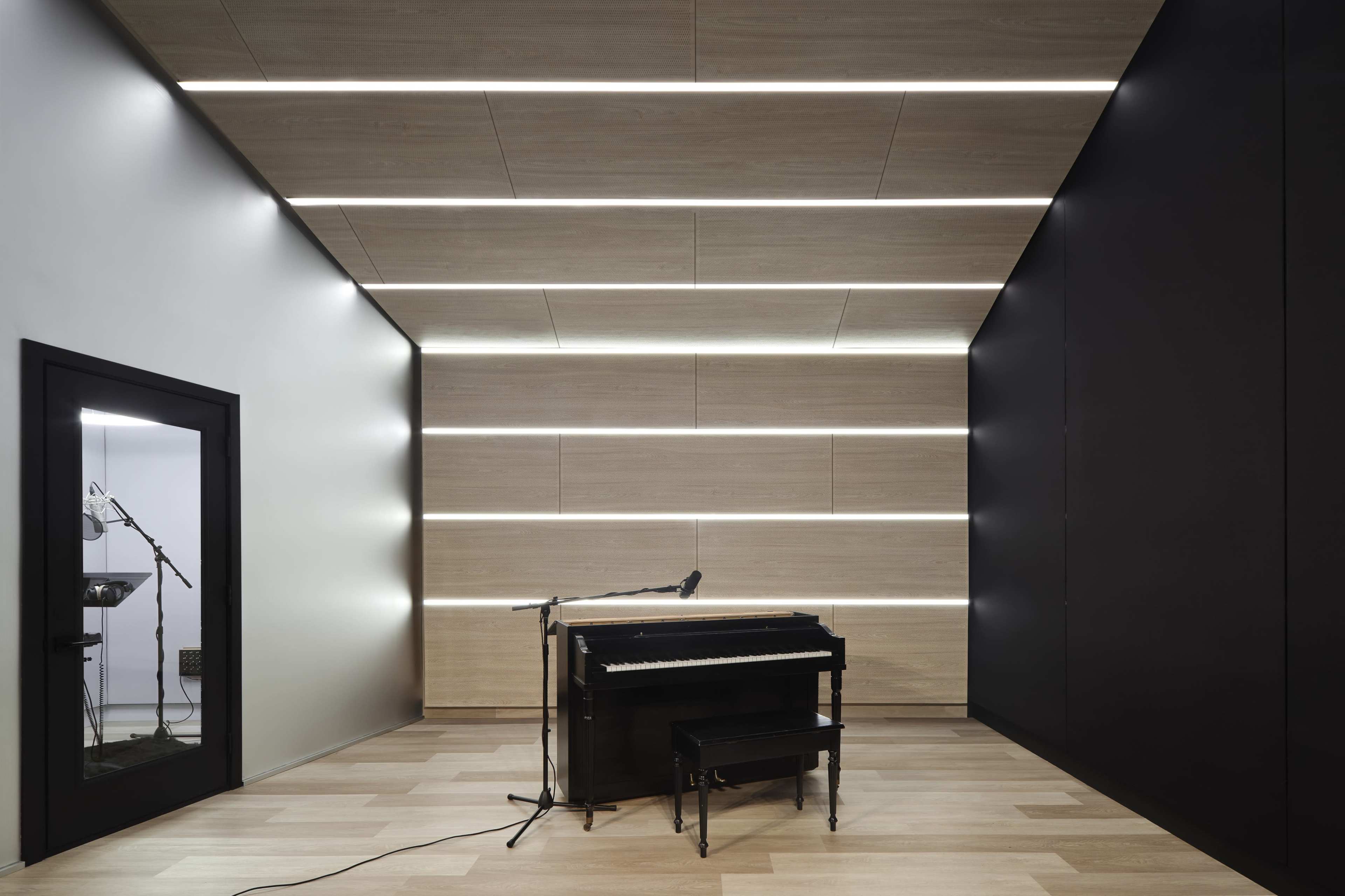 The image shows a minimalist music studio featuring a black upright piano positioned in the center with a microphone nearby, surrounded by wooden walls and strips of modern lighting.