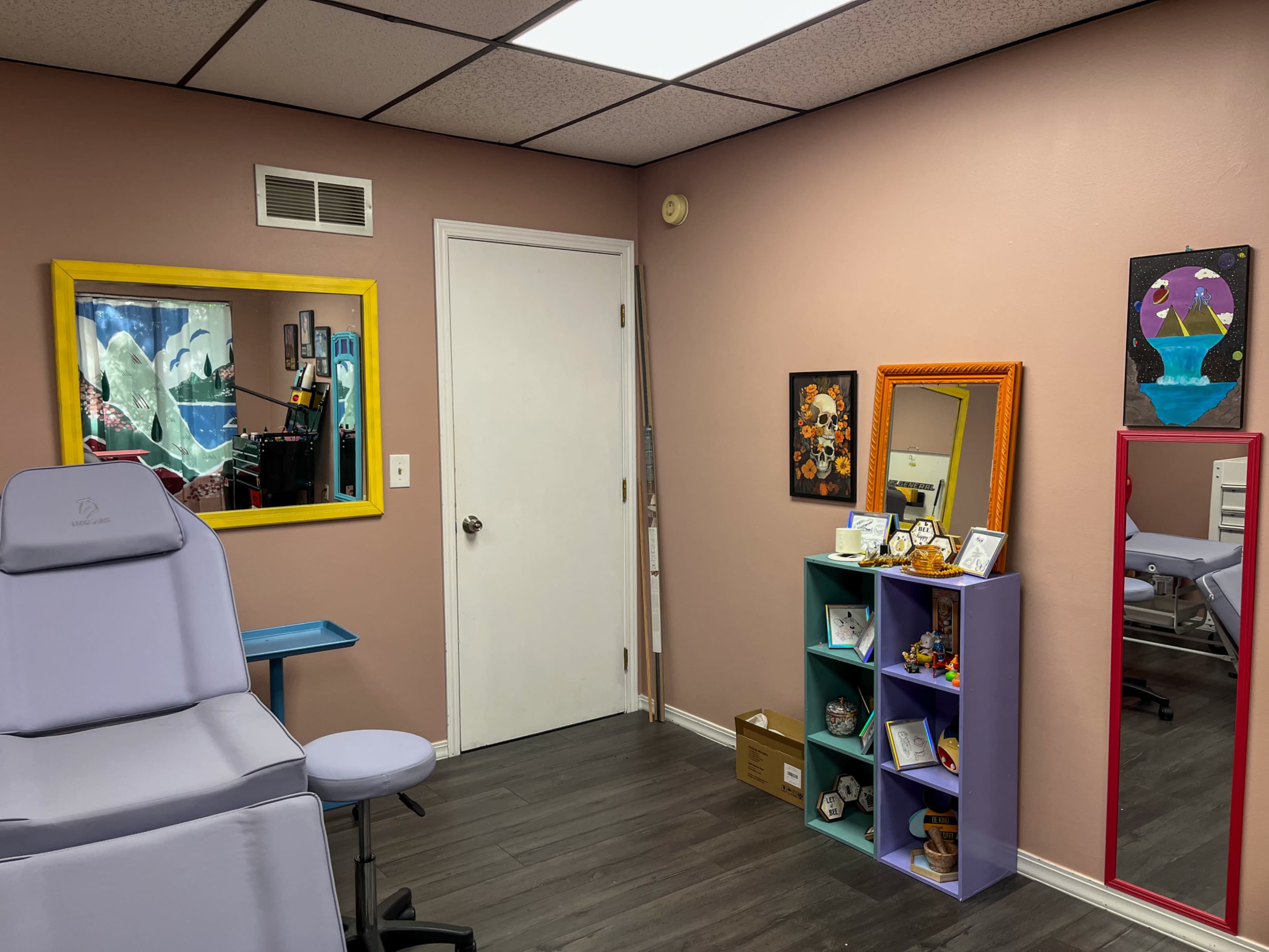 The image shows a small room with pink walls, featuring a treatment chair, a decorative shelf with various items, and mirrors on the walls.