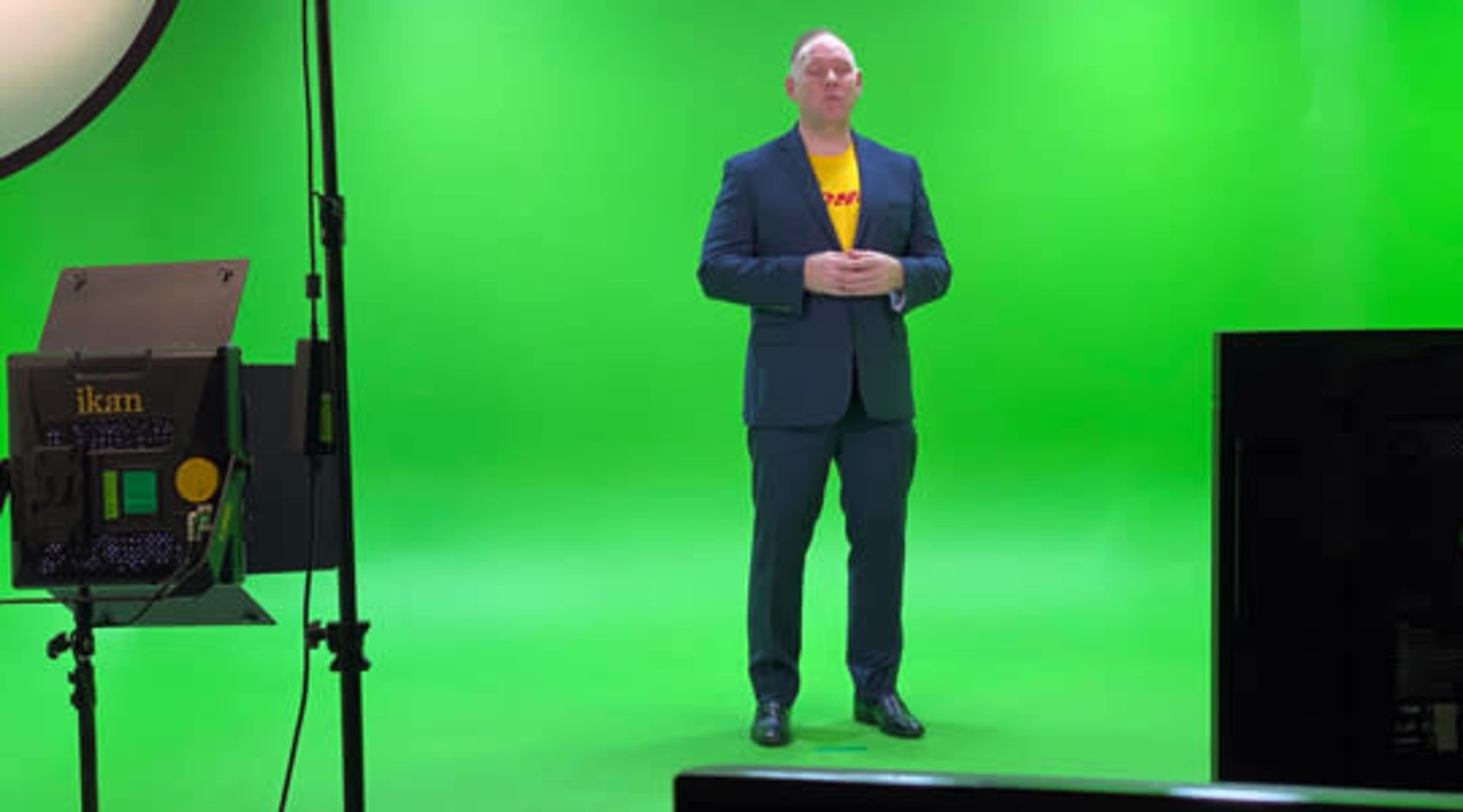 A man in a suit stands on a green screen set with lighting equipment positioned nearby.