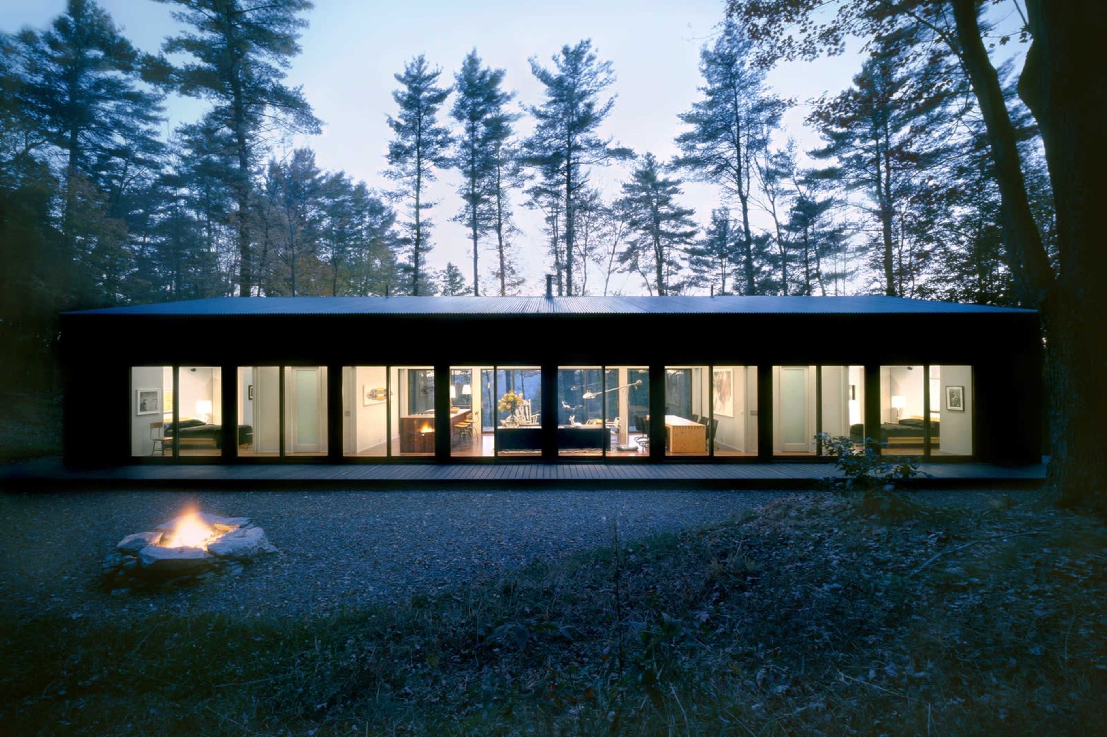 A modern, rectangular house with large windows sits among tall trees, illuminated by interior lights at dusk.