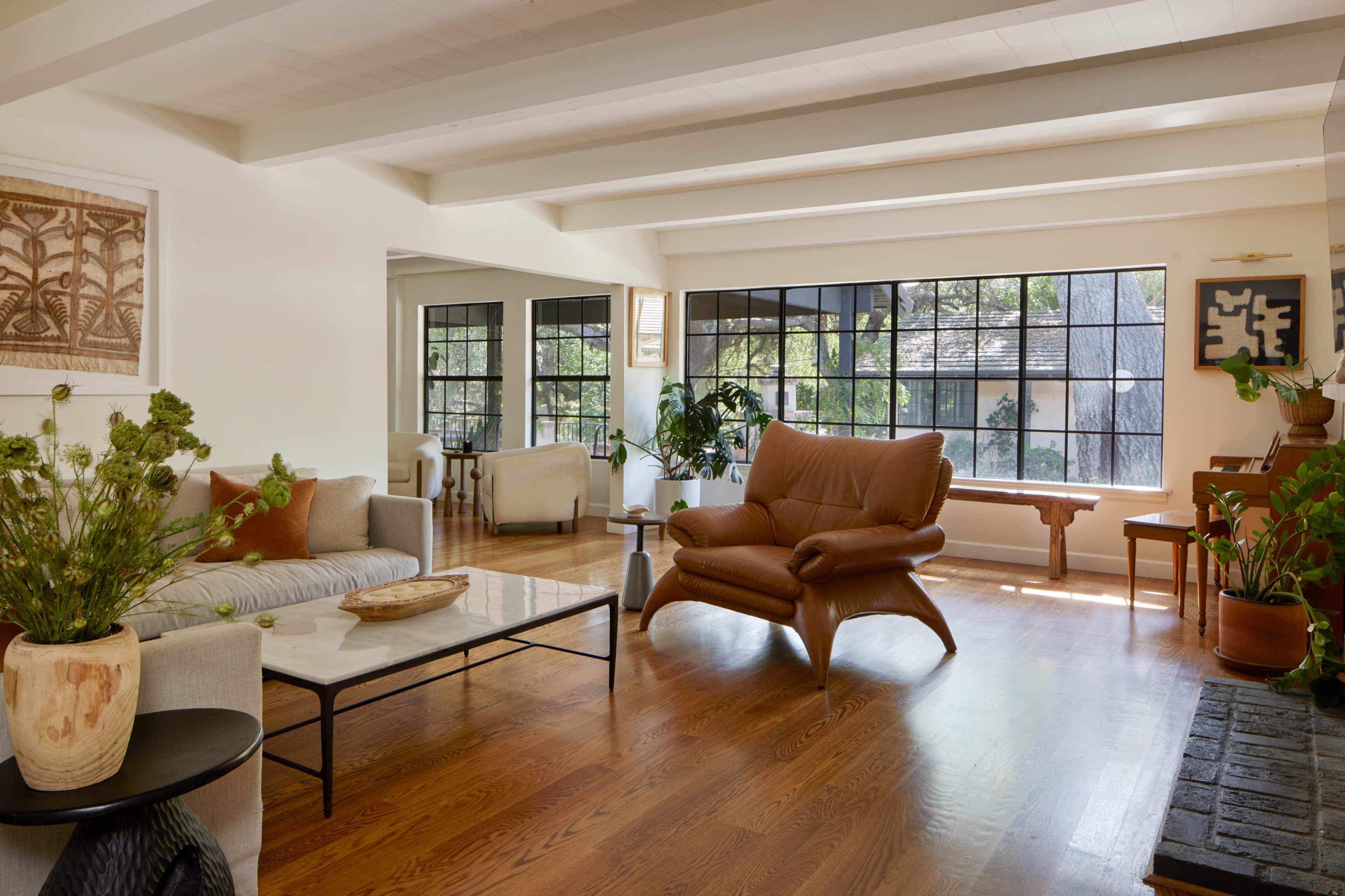 The image depicts a bright, open living room with large windows, wooden floors, and modern furniture, including a brown leather chair and a coffee table.