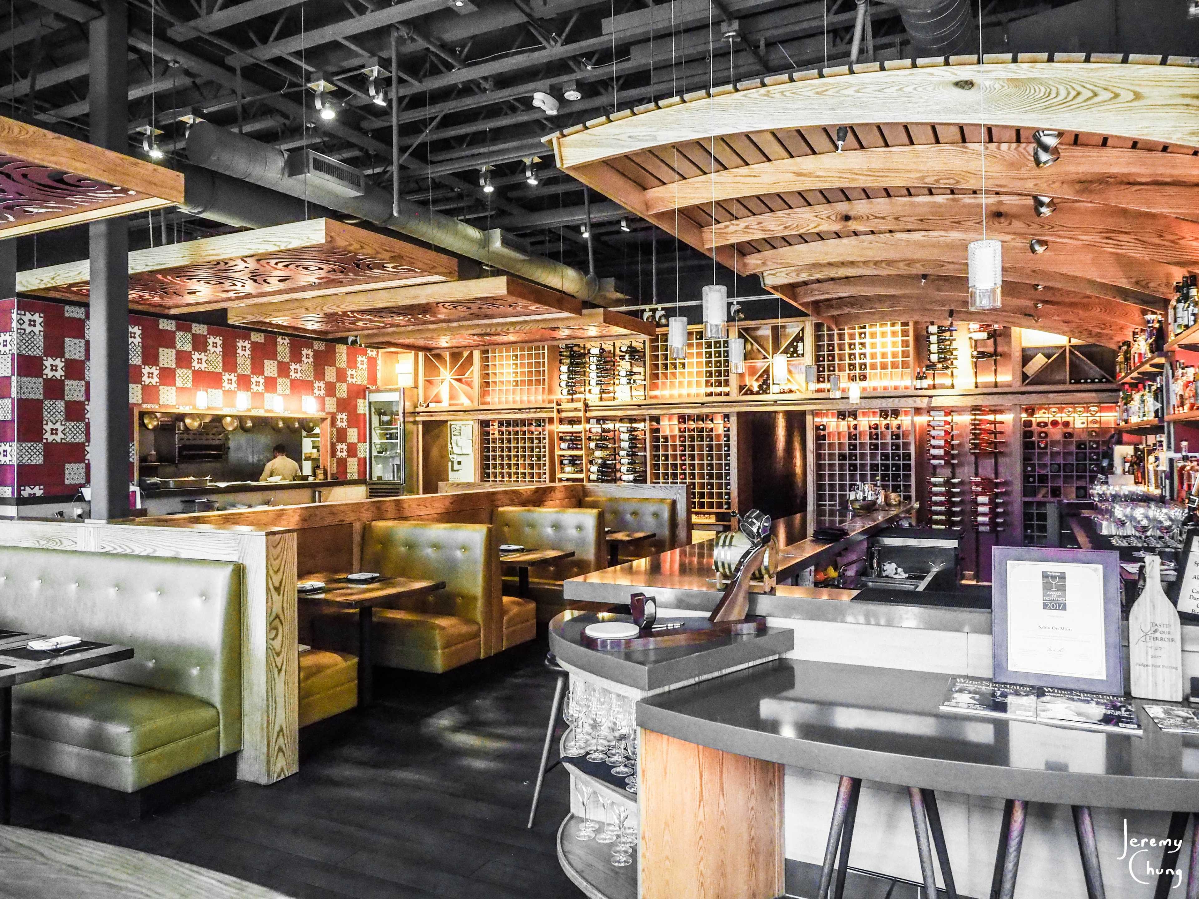 The interior of a modern restaurant featuring wooden ceilings, booths with gray upholstery, and a wine rack displayed on the wall.