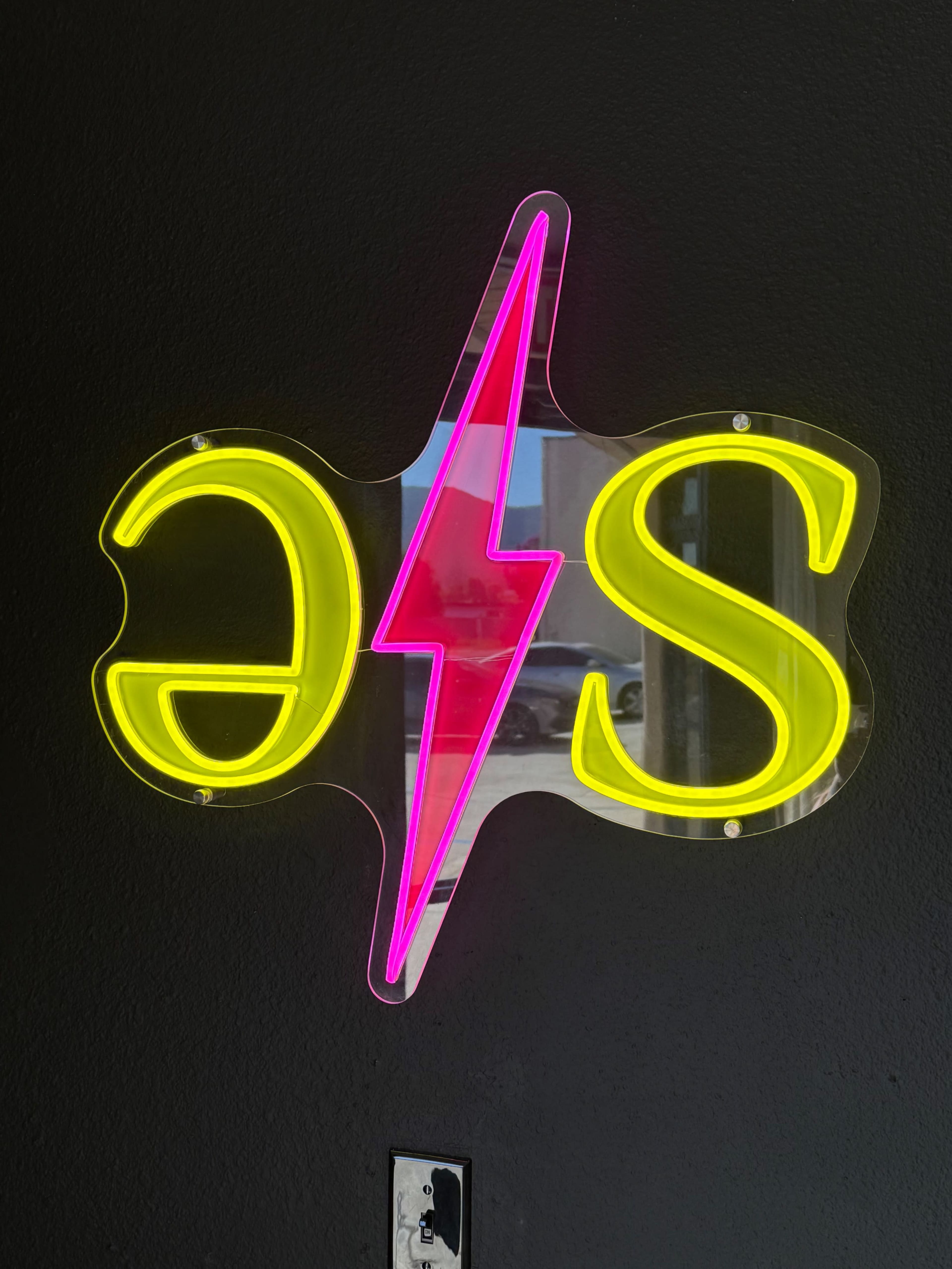 A neon sign displaying the letters "S" and a stylized lightning bolt in pink and yellow colors hangs on a black wall.
