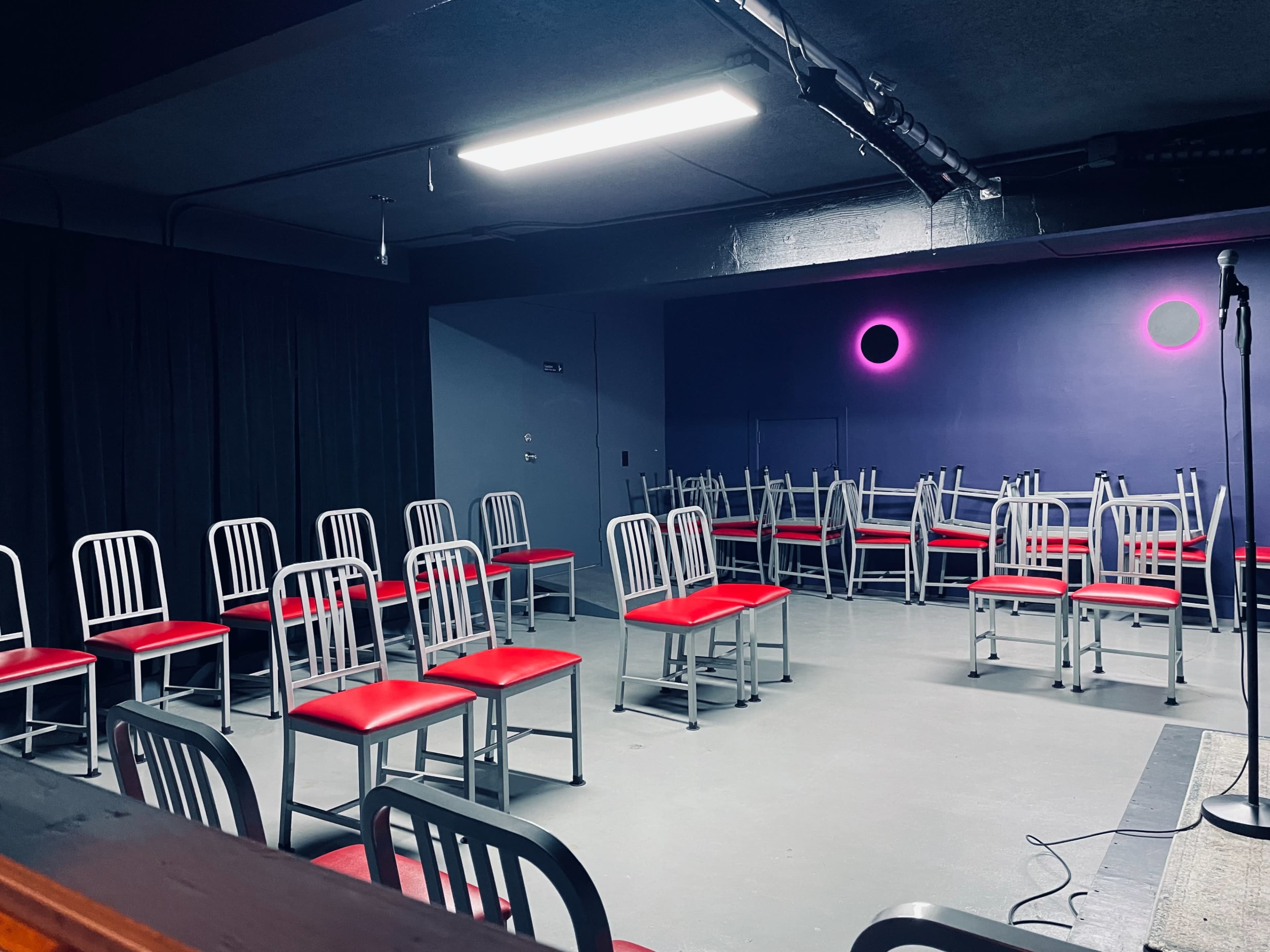 The image shows a dimly lit room with a set of metal chairs arranged around a small stage, featuring a microphone stand and a purple wall with circular lighting.