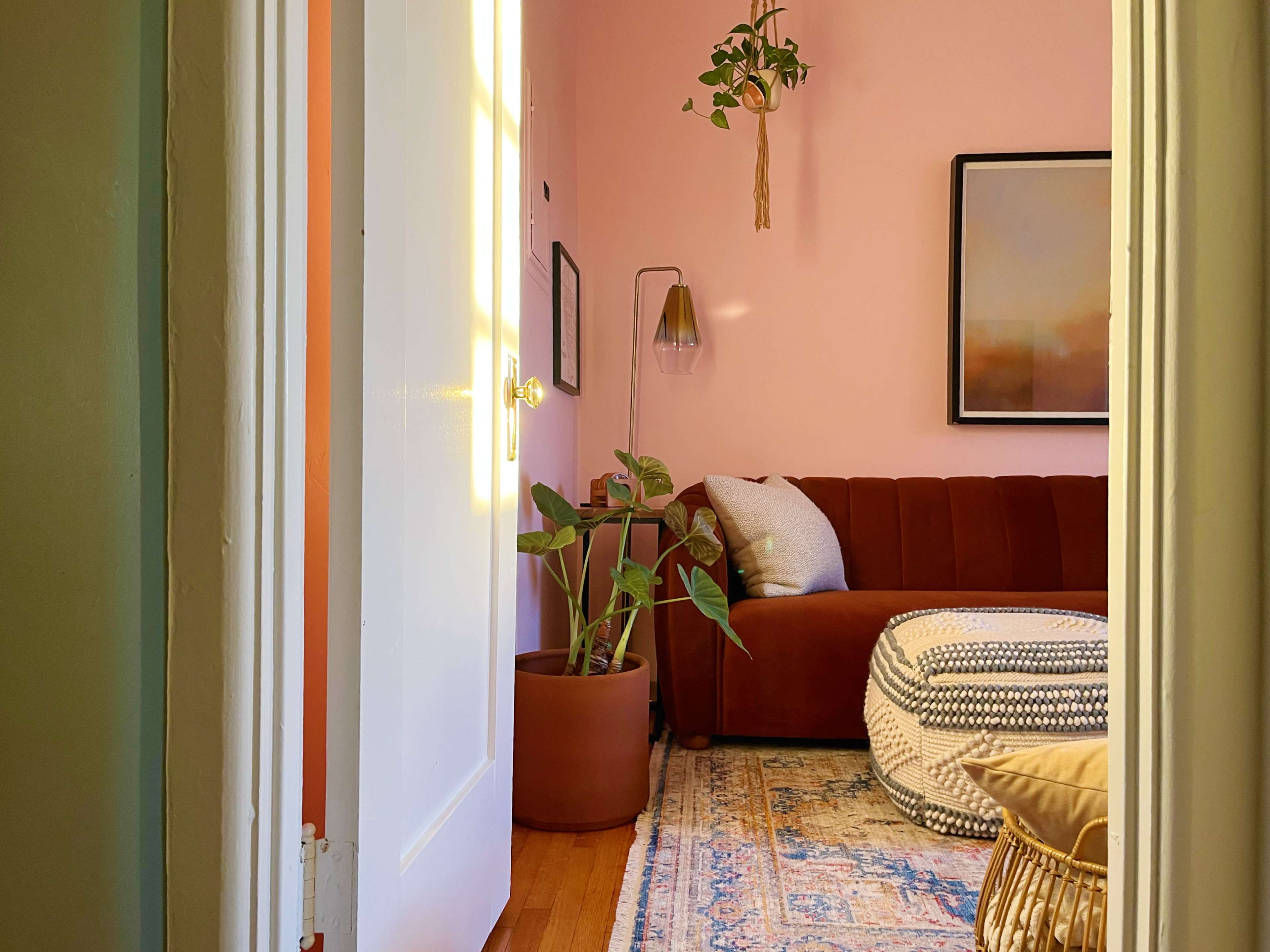 A room with pink walls features a brown sofa, a potted plant, and a framed artwork, visible from an open doorway.