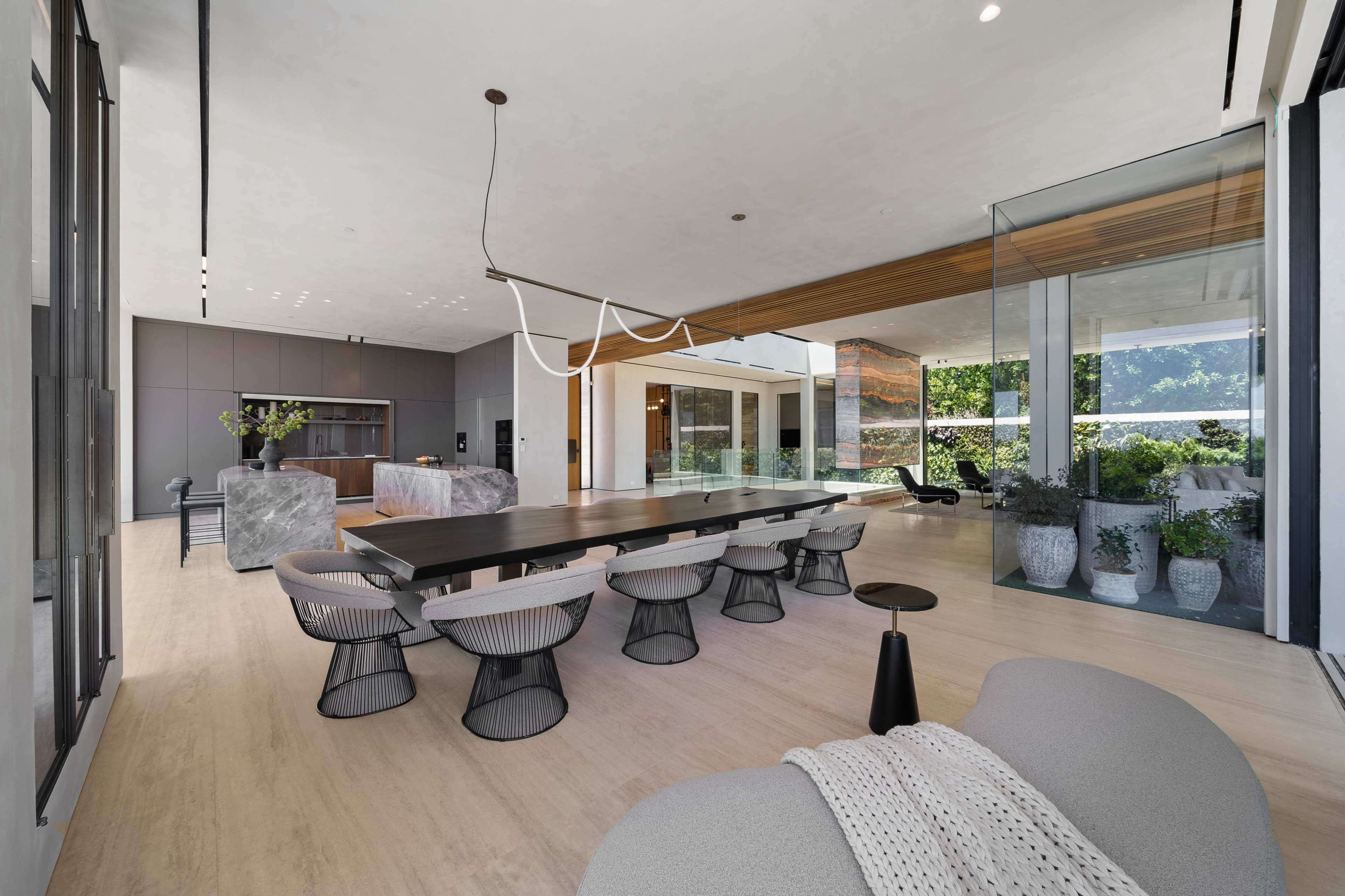 The image shows a modern dining area featuring a long black wooden table surrounded by wire-framed chairs, with large glass windows overlooking an outdoor space.