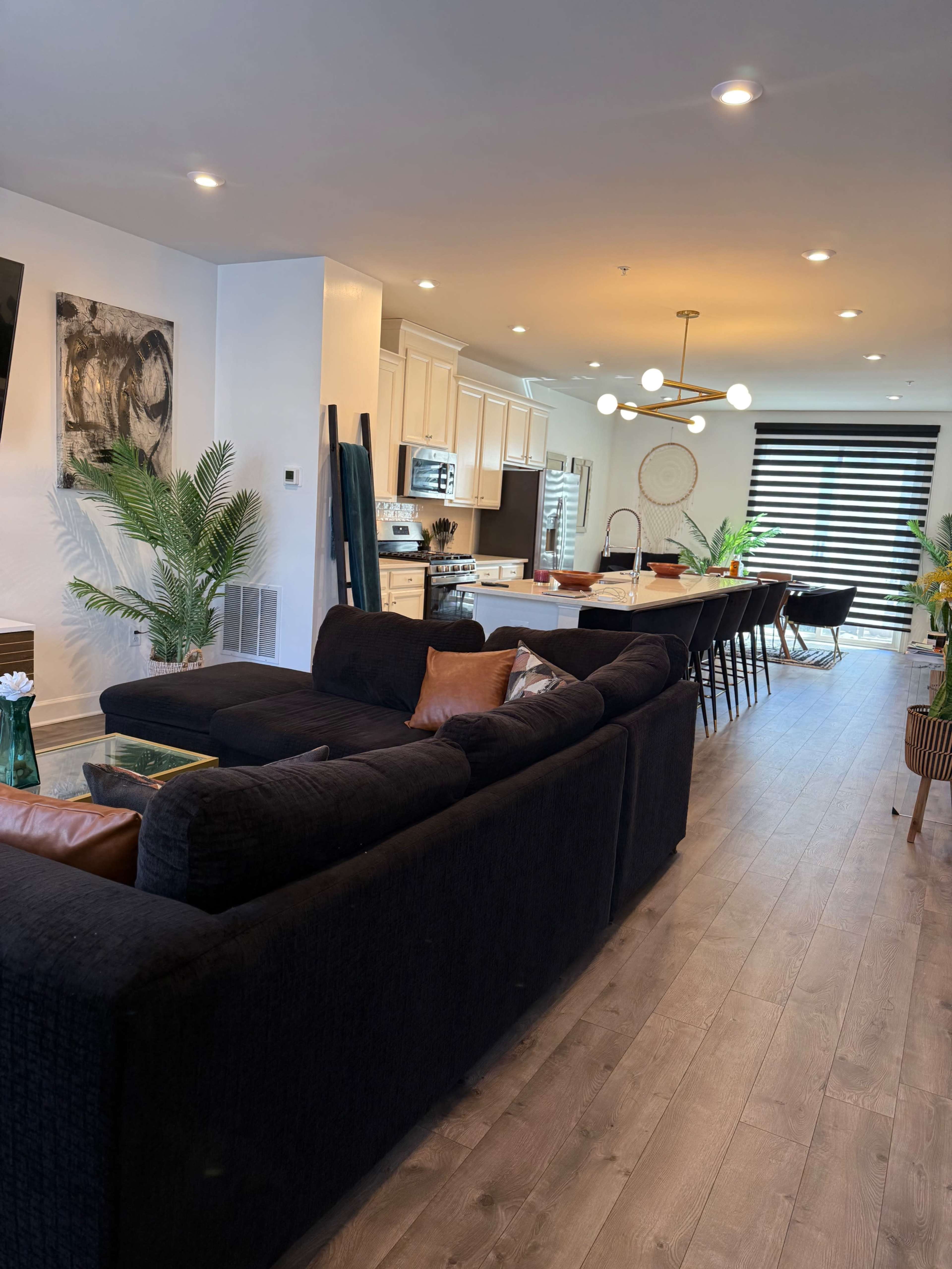The image shows a modern open-concept living space with a dark sectional sofa, a kitchen with white cabinets, and a dining area featuring a long table and decorative plants.