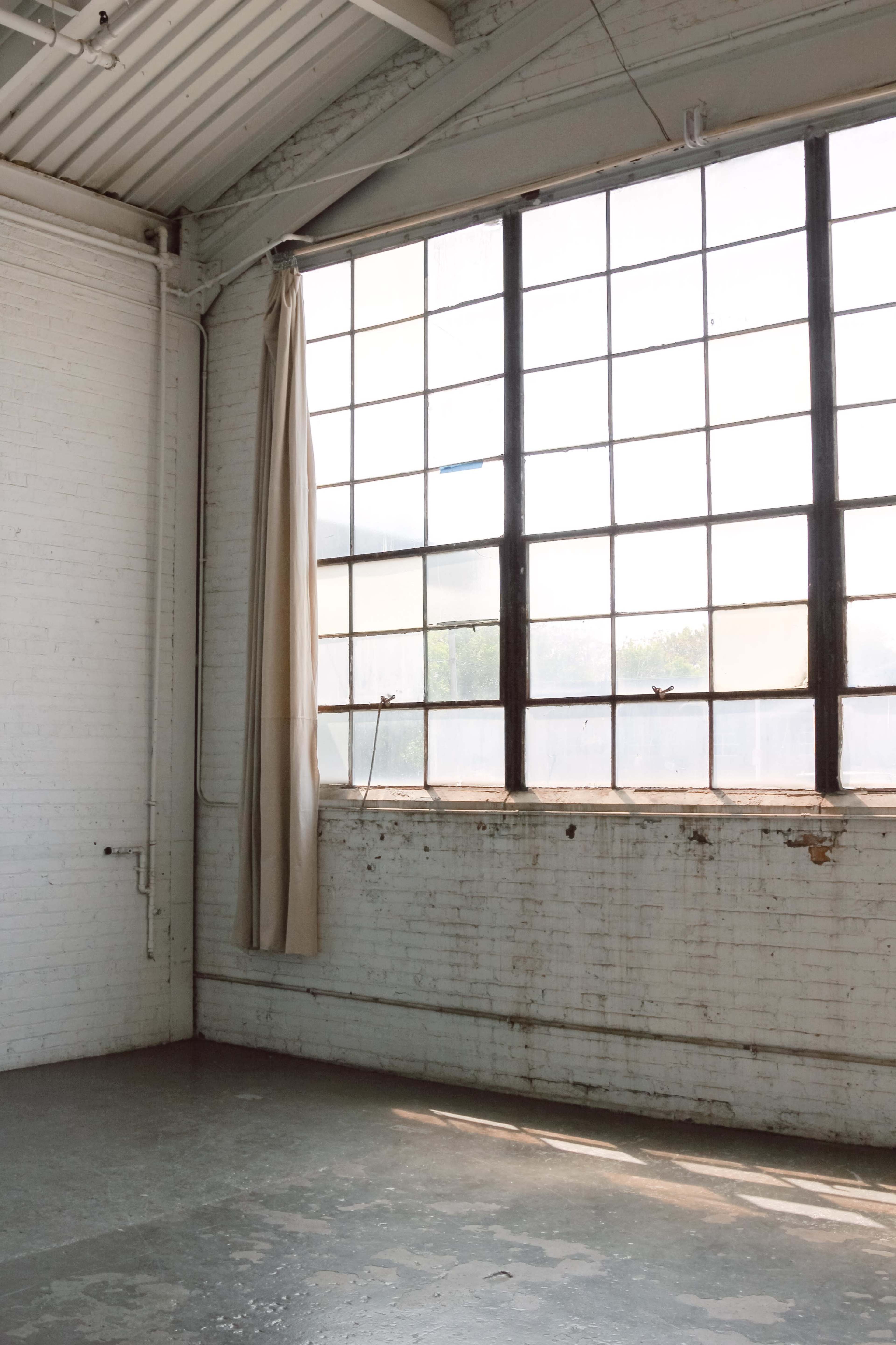 A large industrial window with a curtain is set in a brick wall of an empty room with a concrete floor.