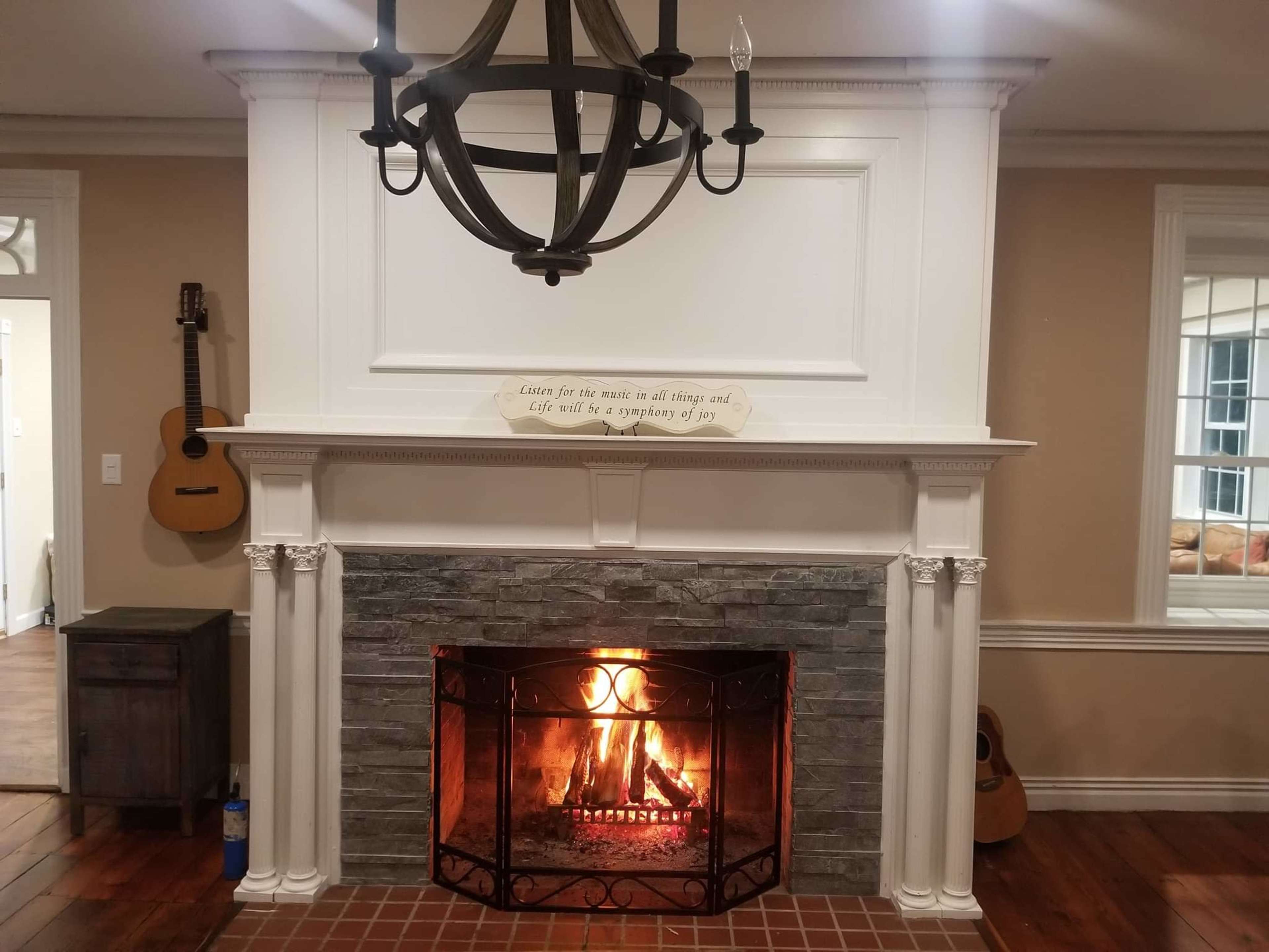 A cozy living room with a stone fireplace, a lit fire inside the fireplace, and a wooden chandelier hanging above.