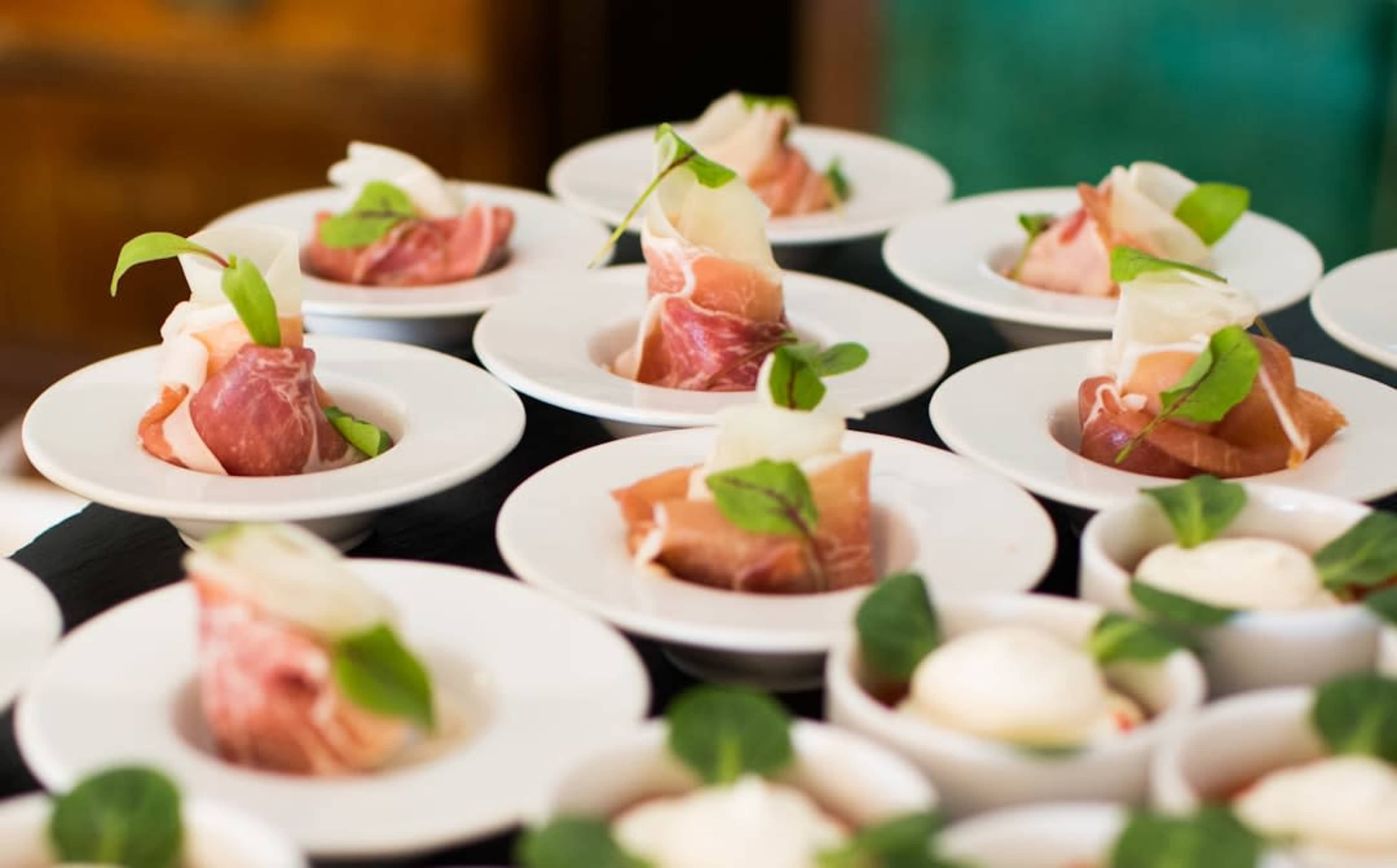 Several small white plates are arranged on a tray, each containing a portion of thinly sliced meat garnished with leafy greens.