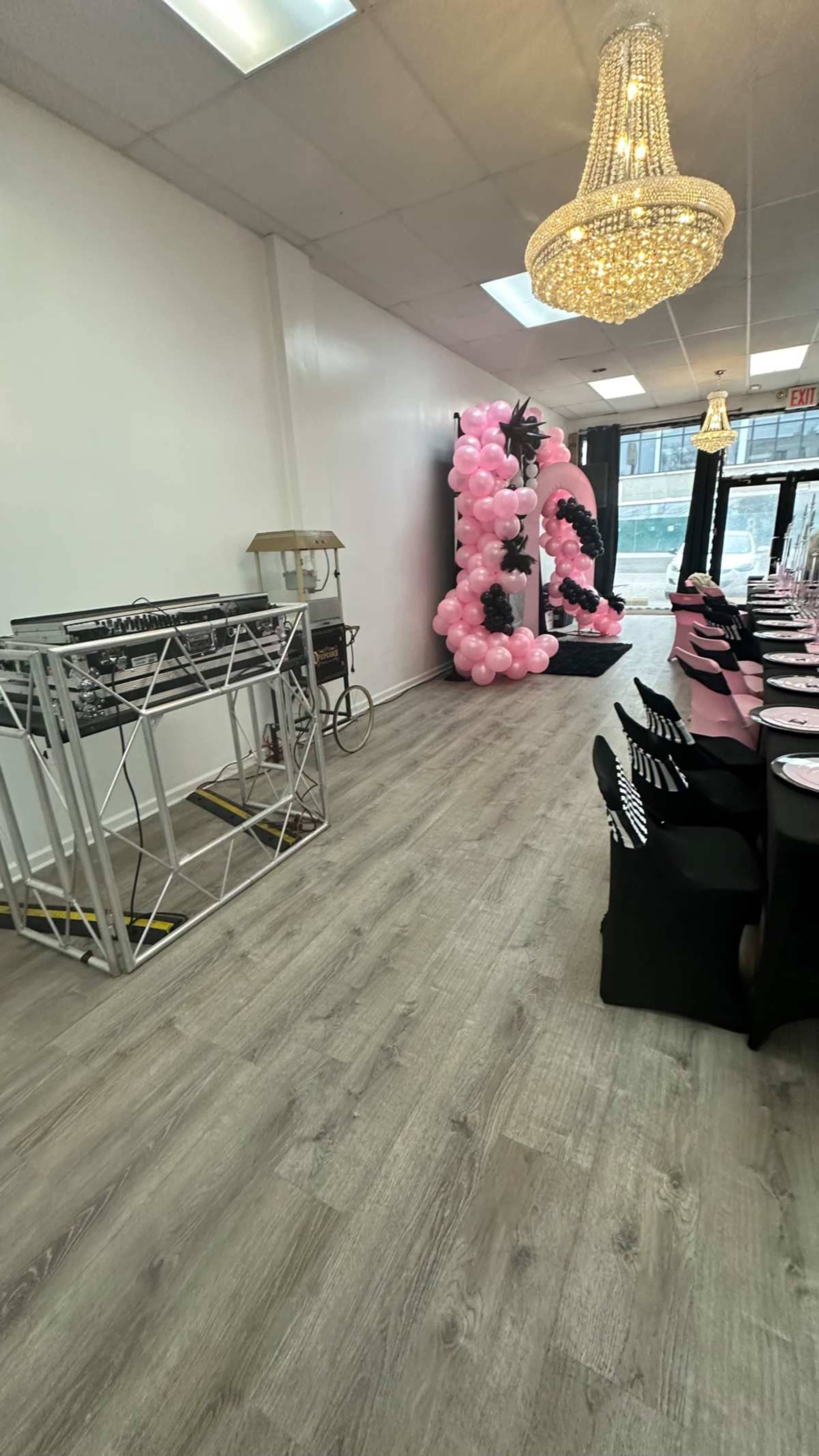 The image shows an indoor event space featuring a line of chairs and tables set for a gathering, with a large pink balloon decoration and a chandelier overhead.