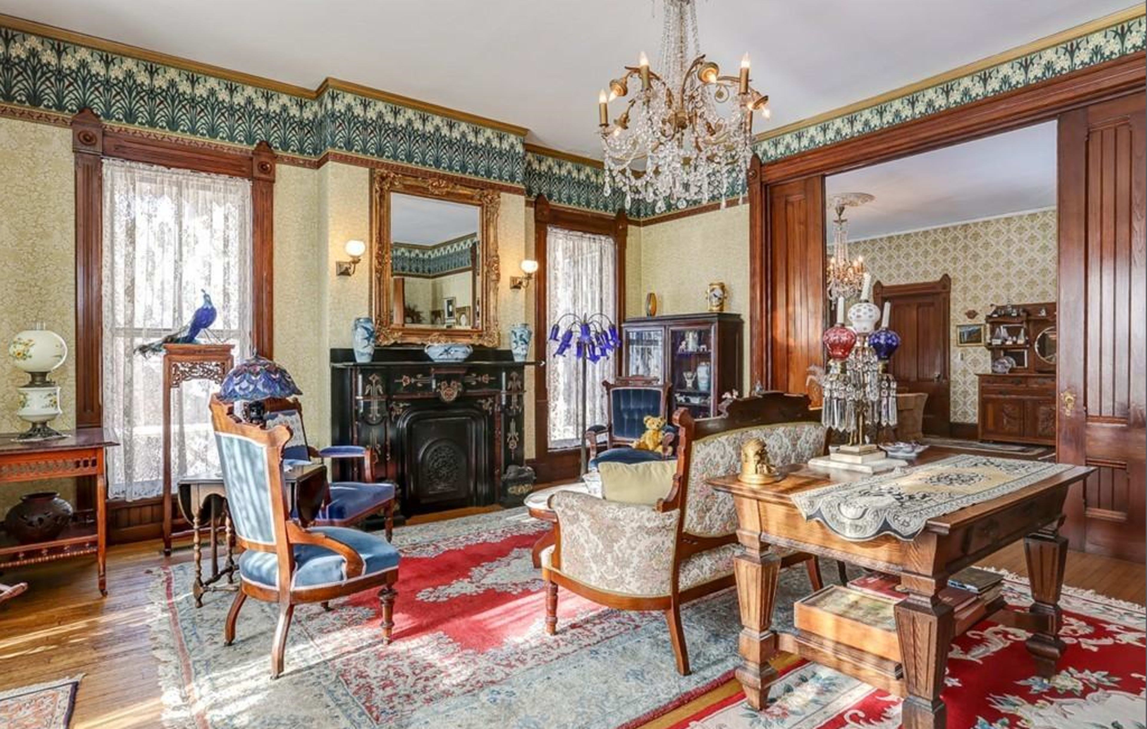 A vintage living room featuring ornate furniture, a chandelier, and decorative wallpaper, with a large rug covering the wooden floor.