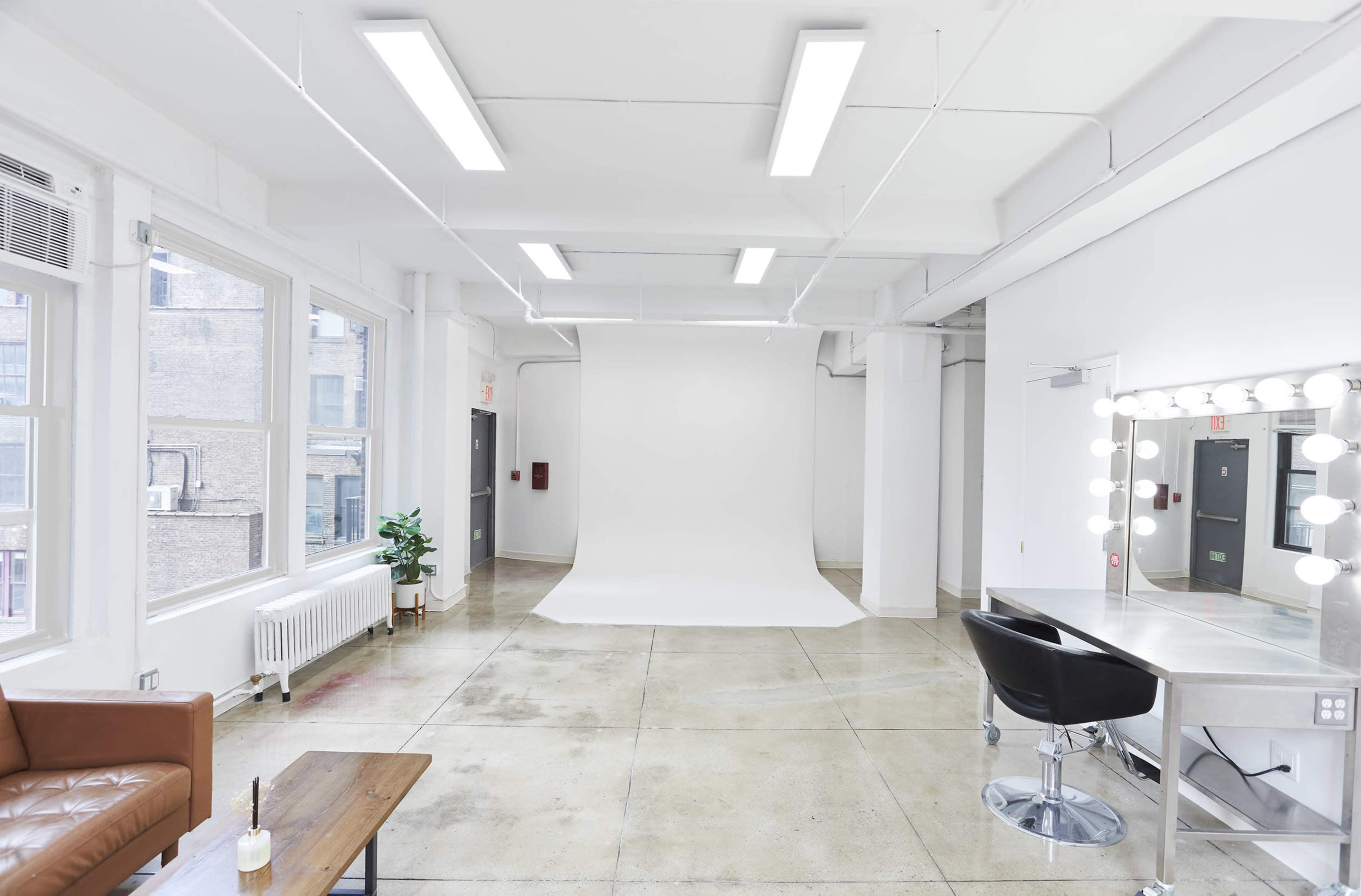 The image shows a bright and spacious photography studio featuring a white backdrop, a mirror with lights, a chair, and a leather couch.