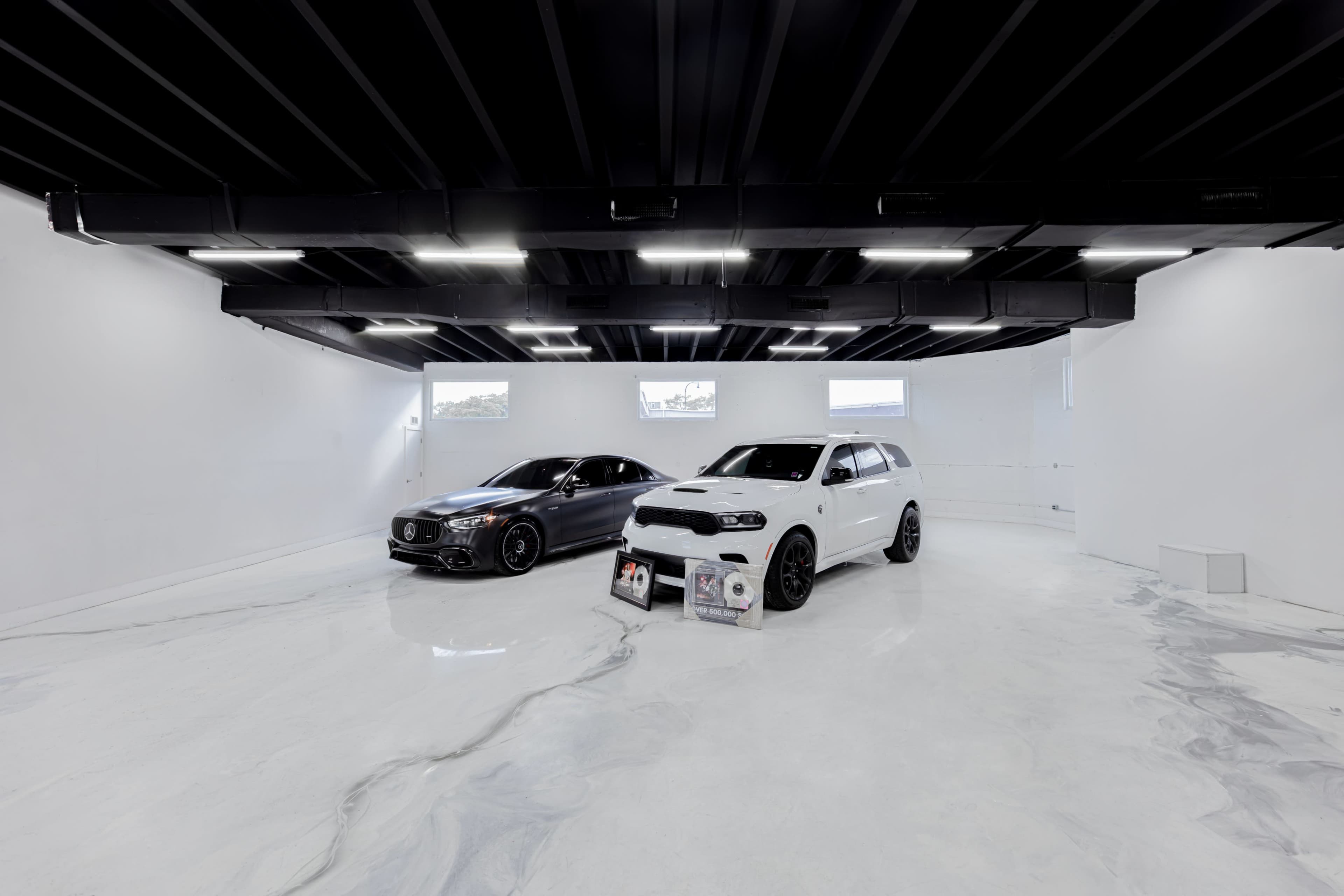 The image shows two cars, a black sedan and a white SUV, parked on a polished white floor in a modern, minimalist showroom with large windows.