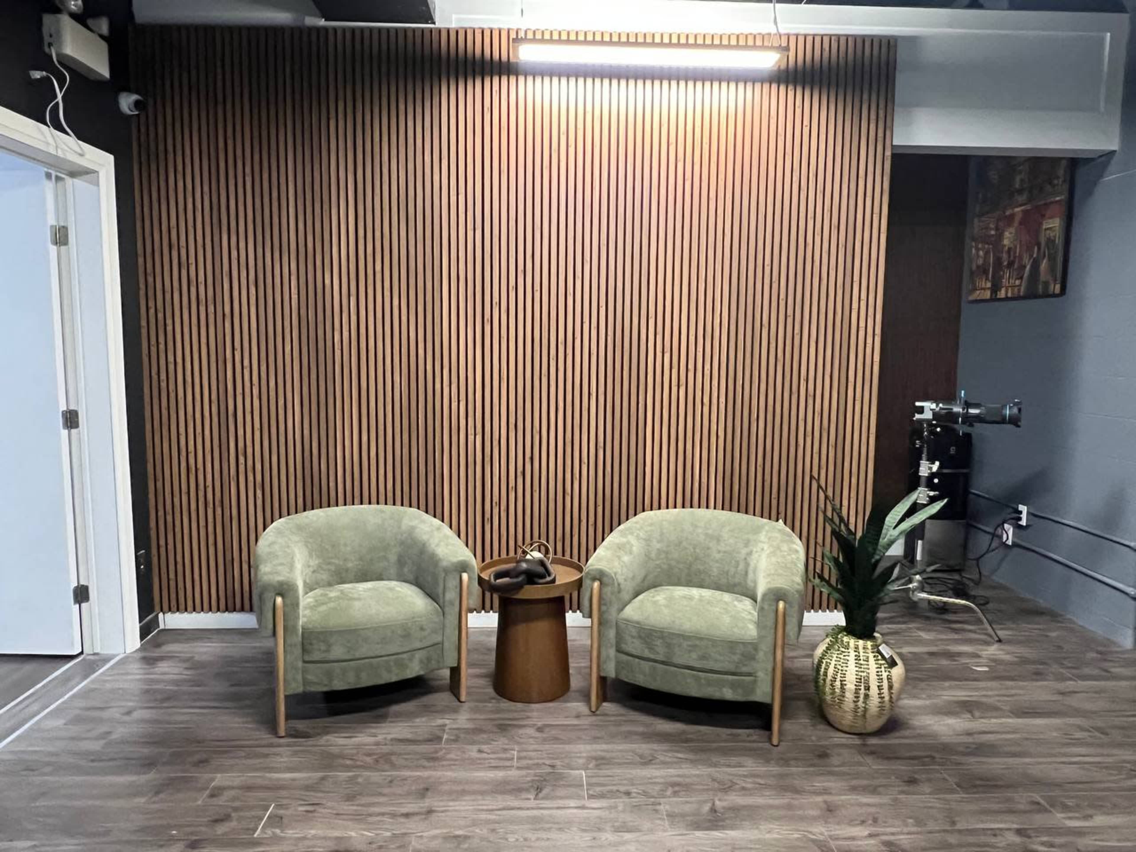 The image shows a modern seating area with two green velvet chairs facing a round wooden table, positioned against a wooden slatted wall.