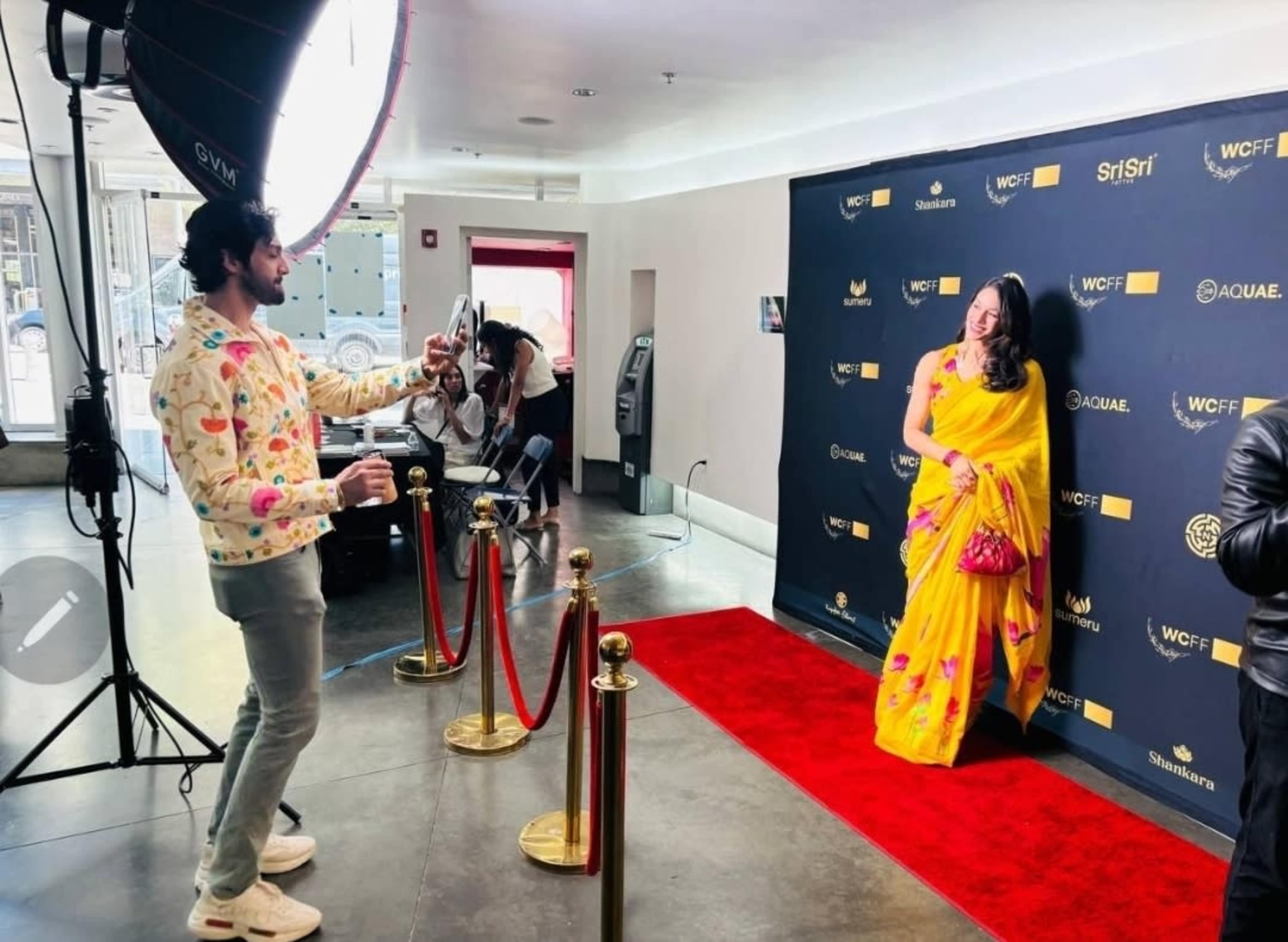 A woman in a yellow saree poses on a red carpet while a man takes her photo in front of a branded backdrop.