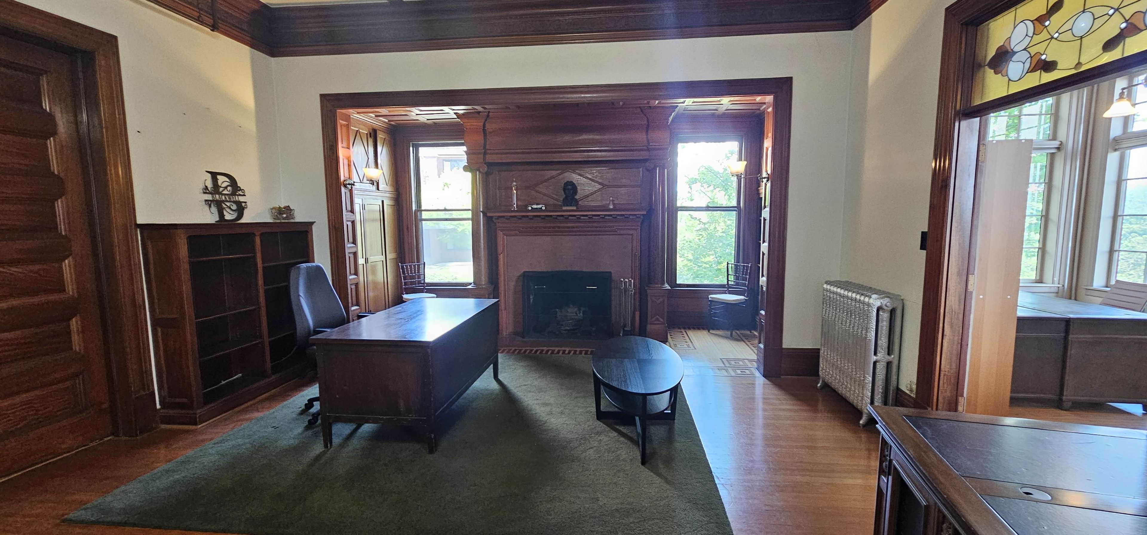 The image shows a well-lit office space with a wooden desk, a chair, and a fireplace, surrounded by windows that provide outdoor views.