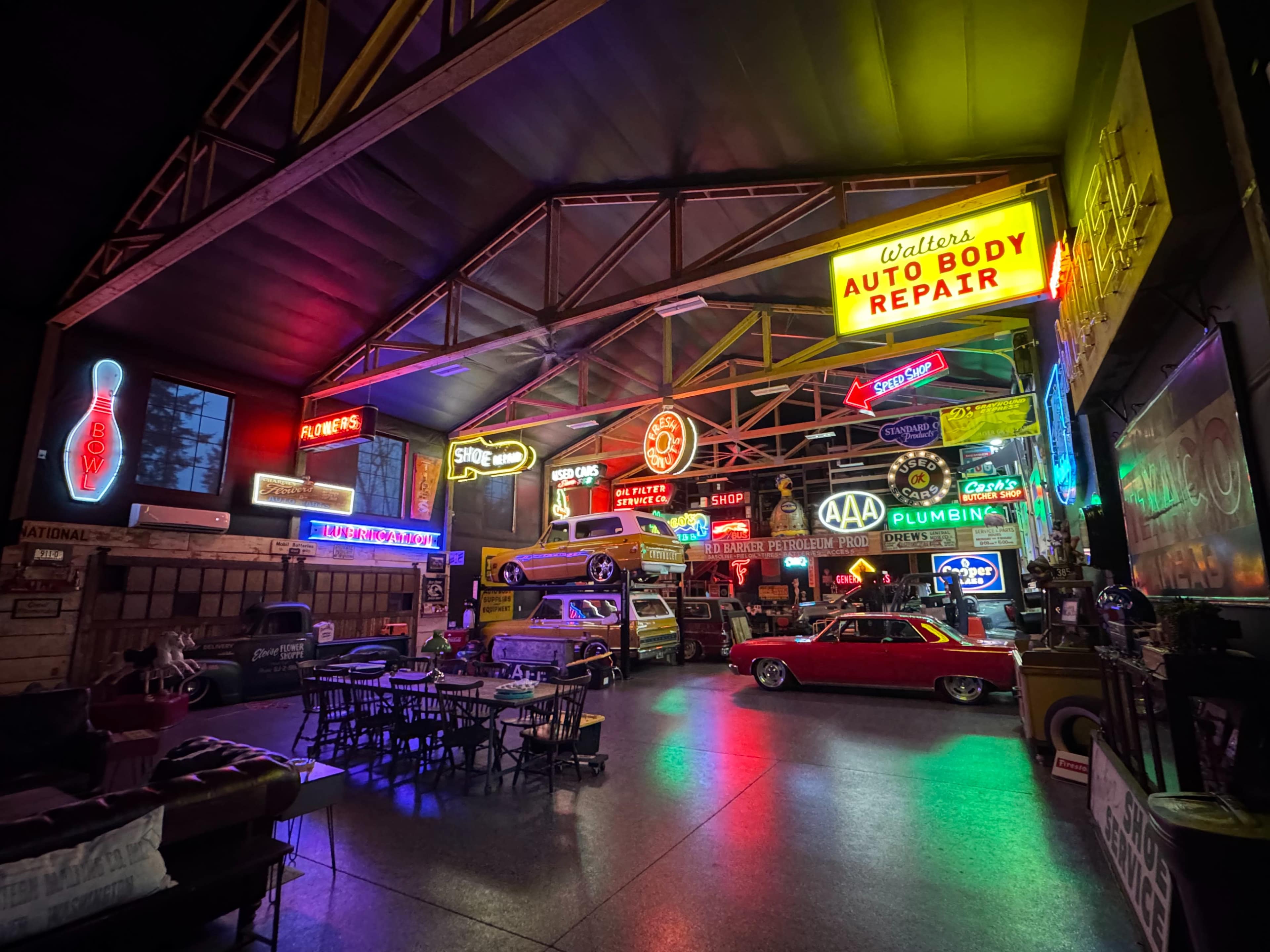 The image shows a retro-themed garage or auto repair shop illuminated by various neon signs, featuring vintage cars and a dining area with tables and chairs.