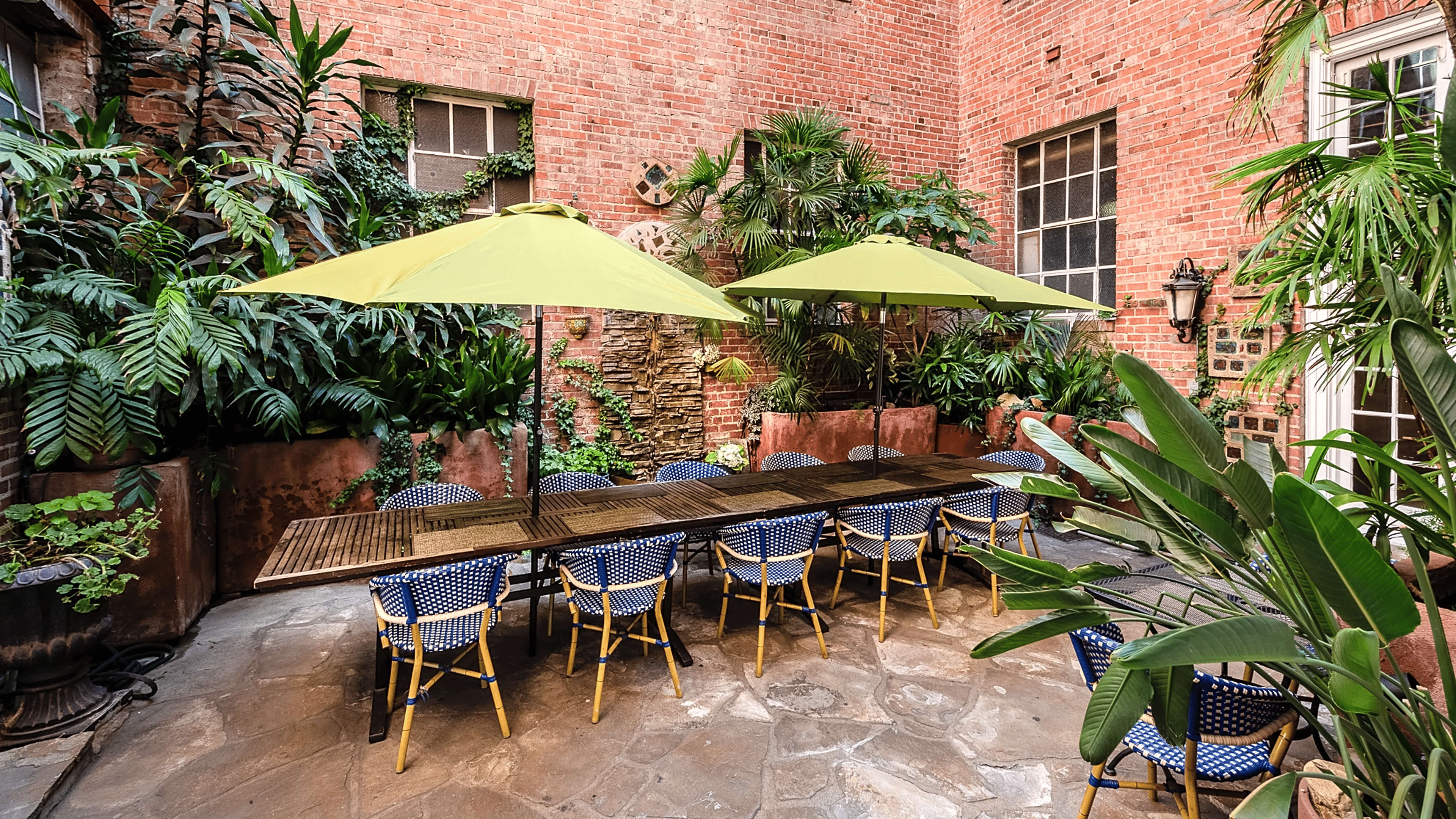 A courtyard features a long wooden table surrounded by blue and white chairs, with two large green umbrellas and lush potted plants against a brick wall.