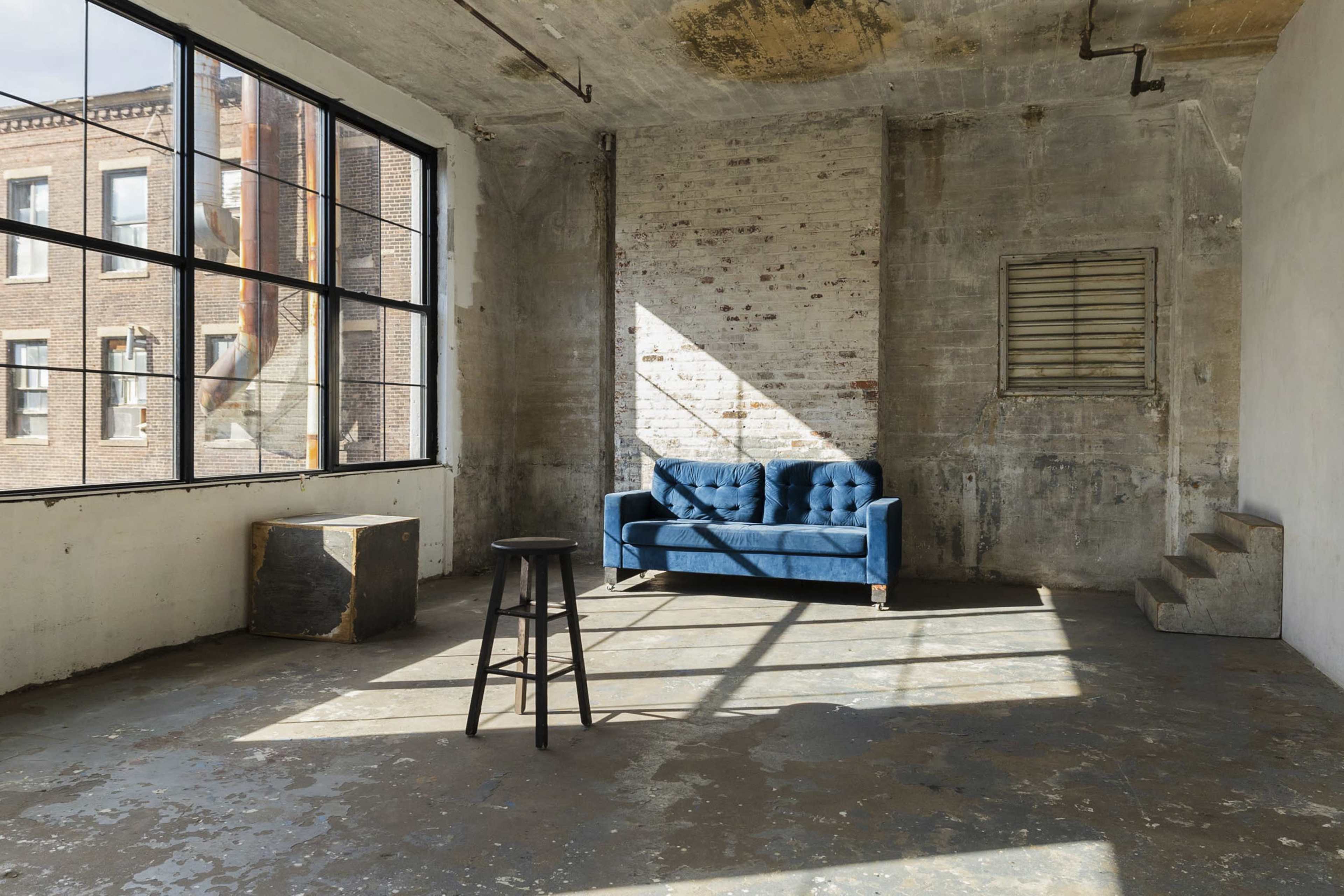 A blue tufted sofa is positioned against a wall in an industrial-style room with large windows and exposed brick.