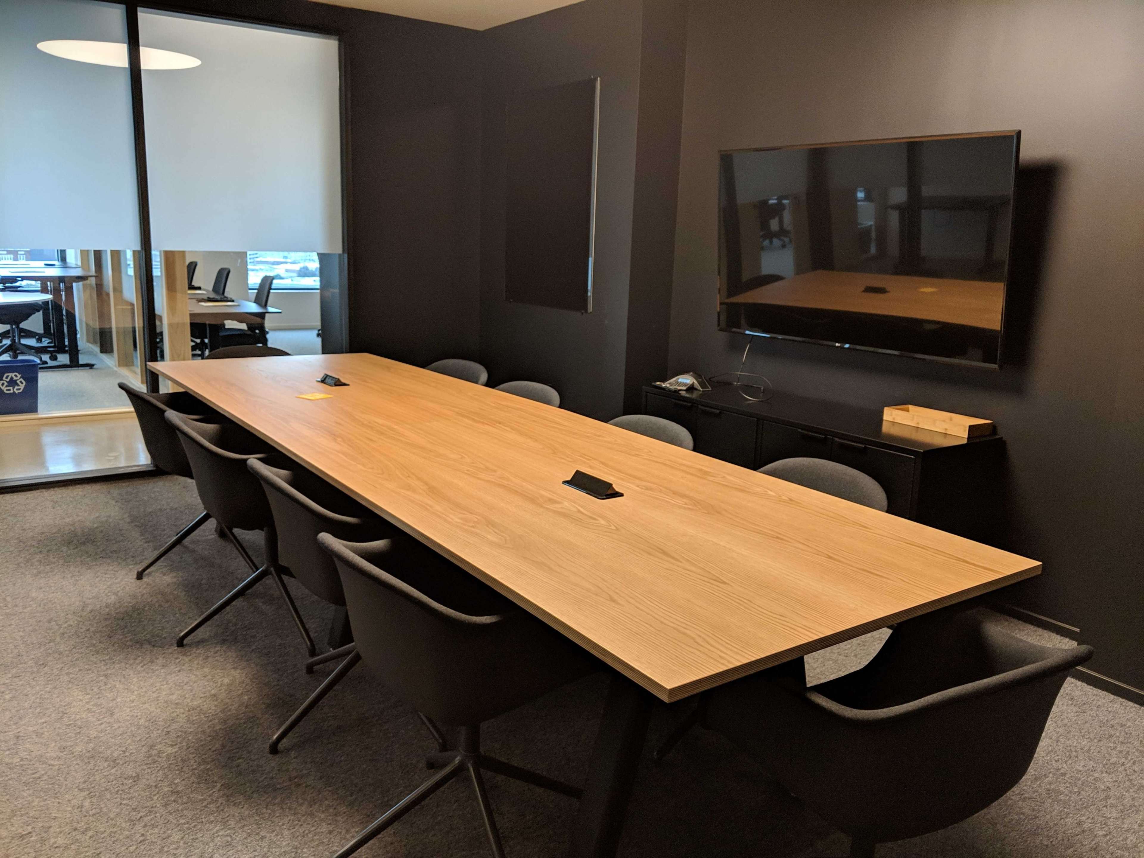 A large wooden conference table with black chairs is positioned in a dark conference room, adjacent to a glass-walled area with additional meeting space.