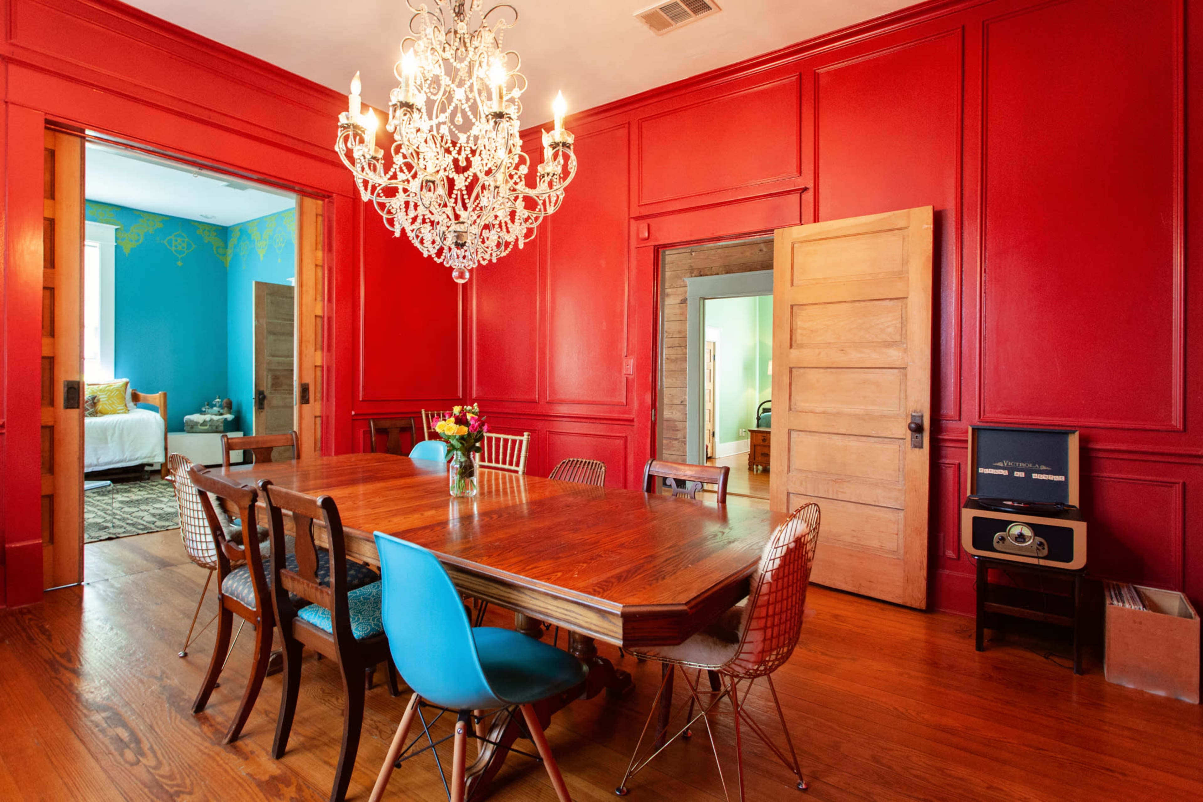 A large wooden dining table is surrounded by a mix of chairs in a room with red walls and a chandelier, with doorways leading to adjacent rooms painted in blue.