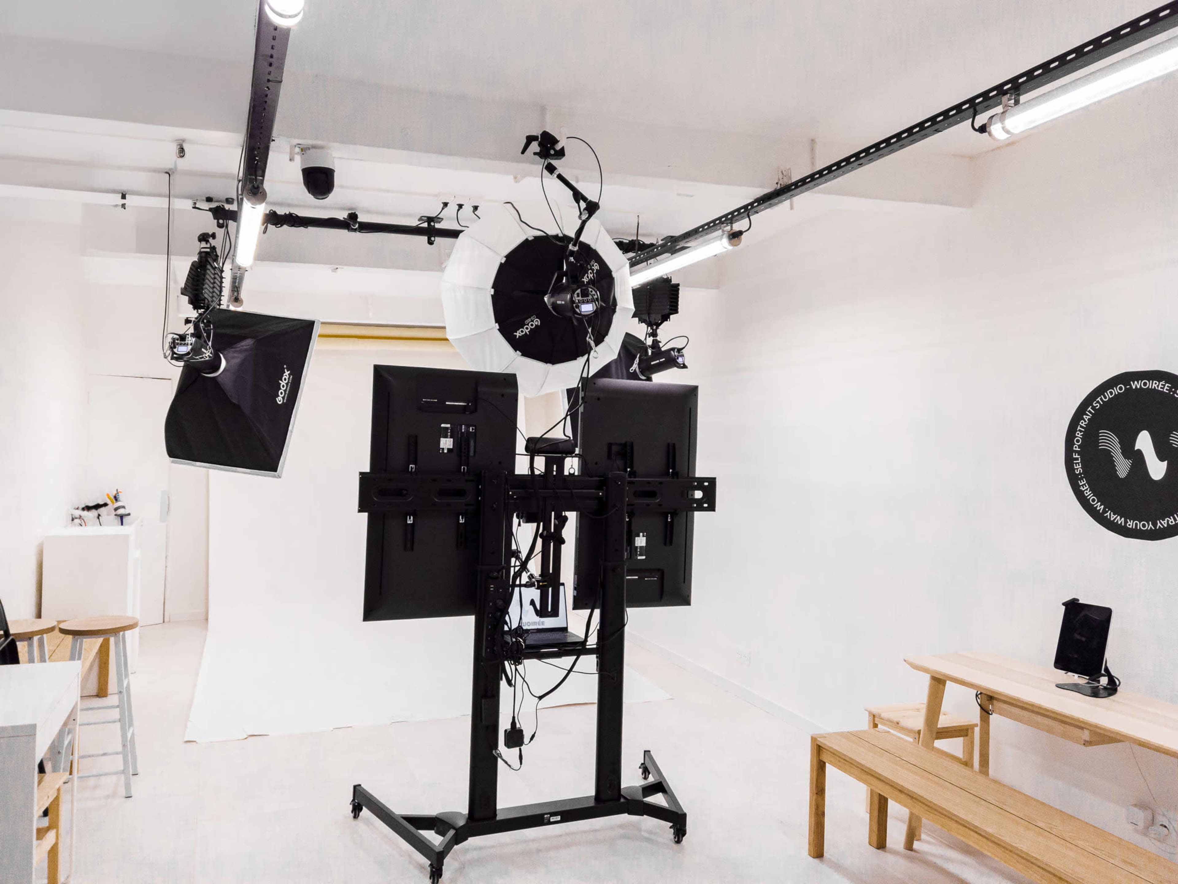 The image shows a well-lit photo studio equipped with multiple light sources, a backdrop, and a dual-screen monitor setup on a movable stand.