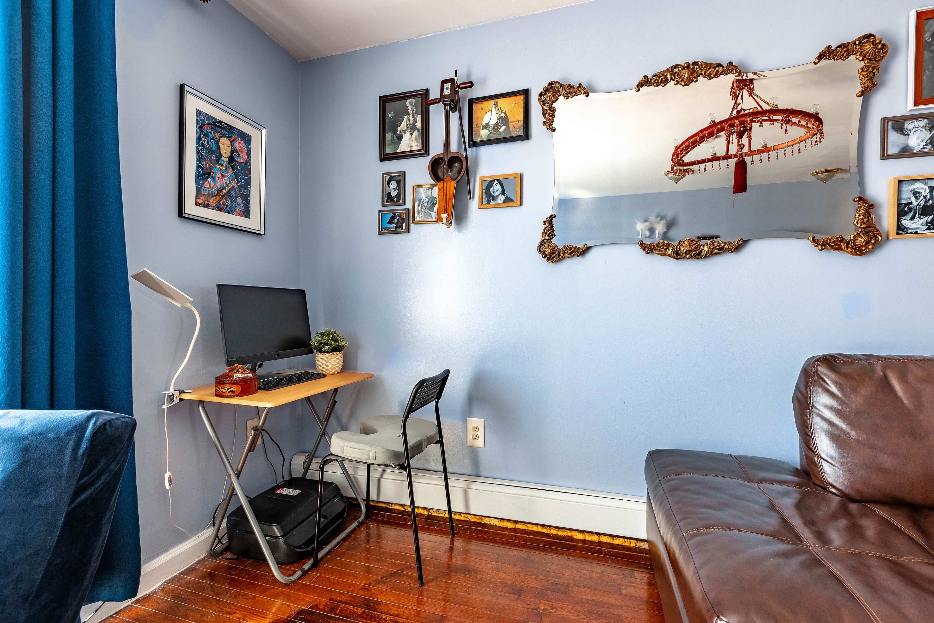 A small room features a desk with a computer, a chair, and various framed photographs on the wall, along with a large mirror and a couch.