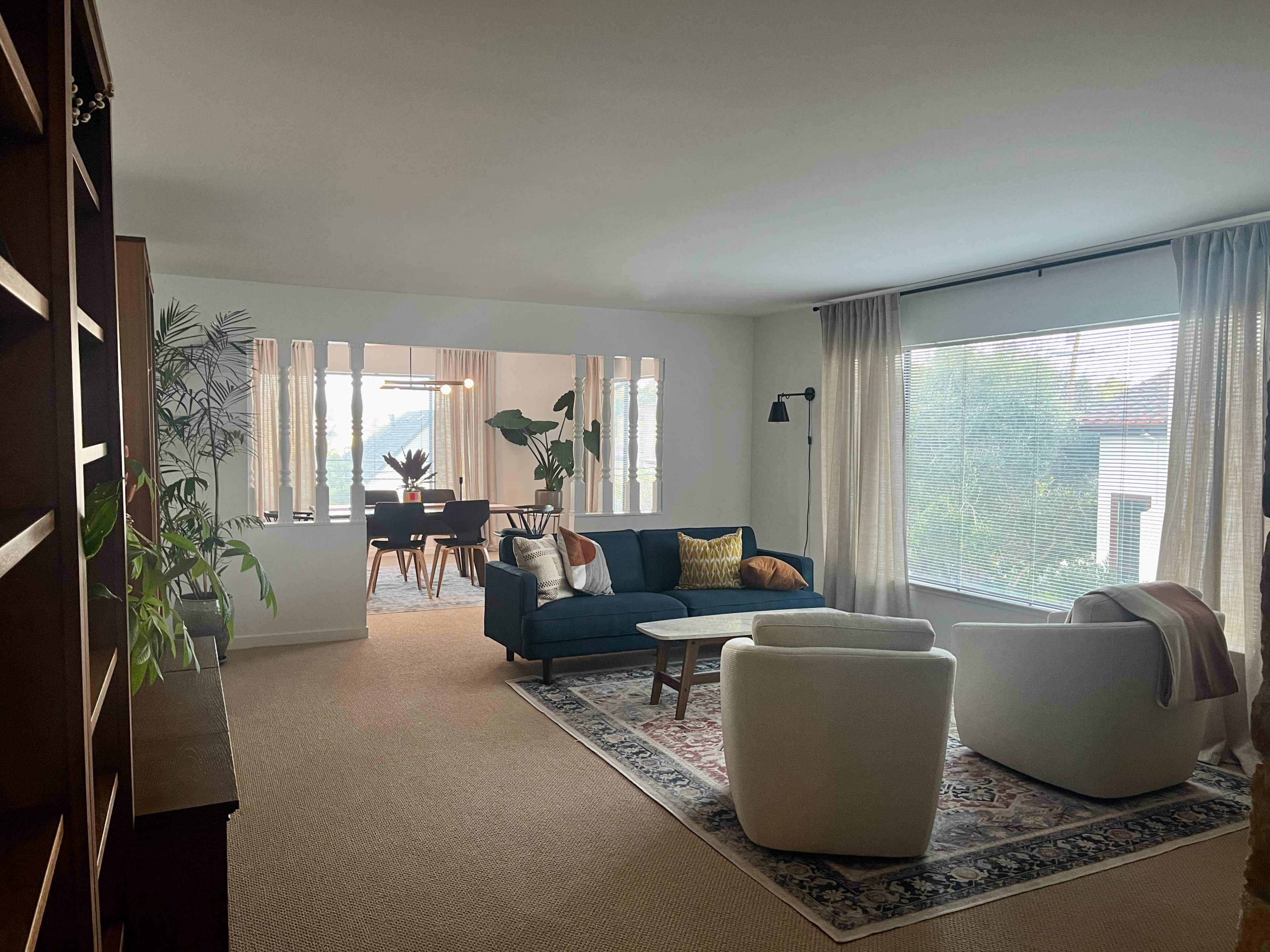 A modern living room featuring a blue couch, two white chairs, a patterned rug, and a dining area visible through an archway with large windows.