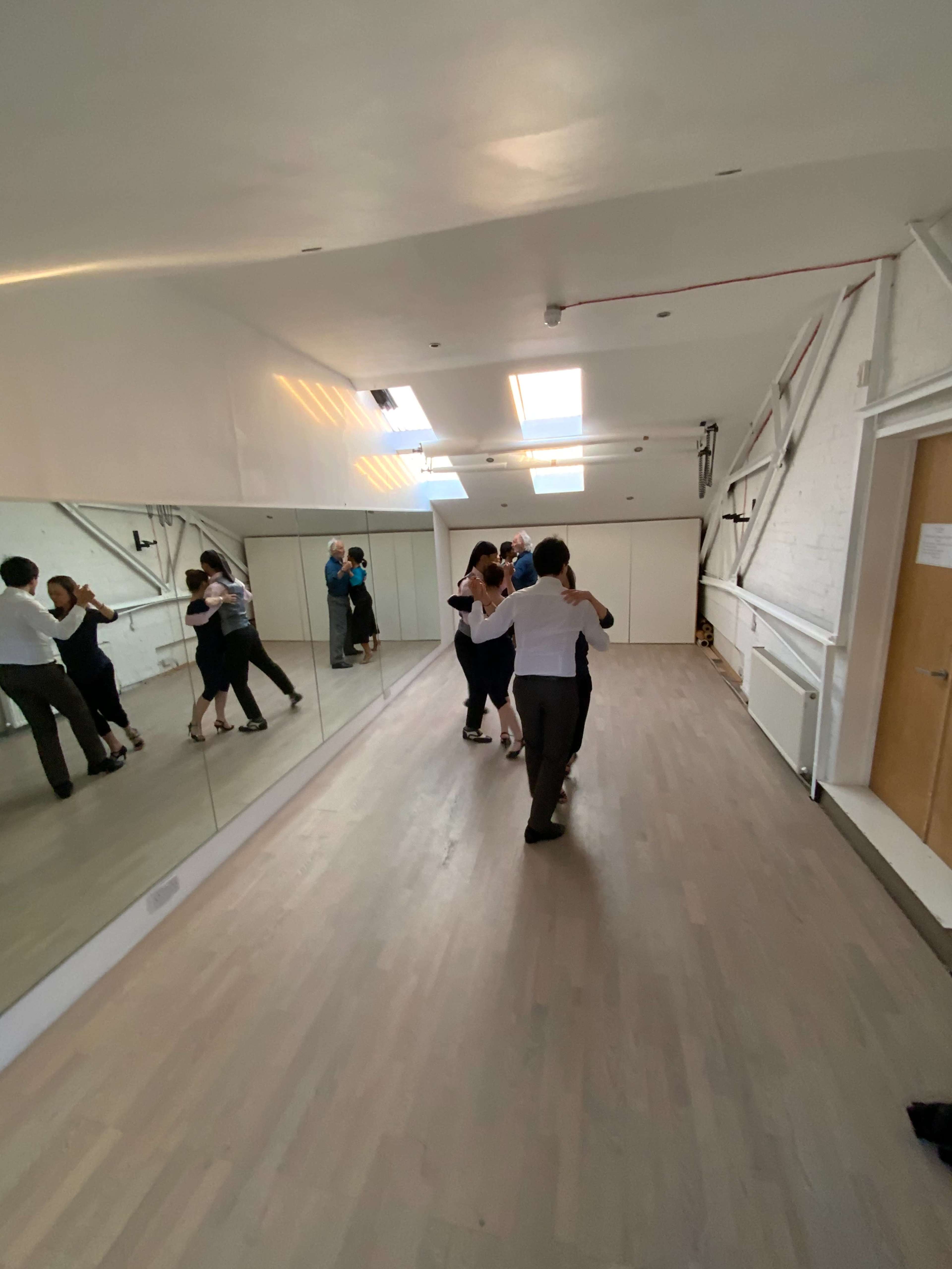 A dance studio features couples practicing in front of a large mirror, with wooden flooring and skylights above.