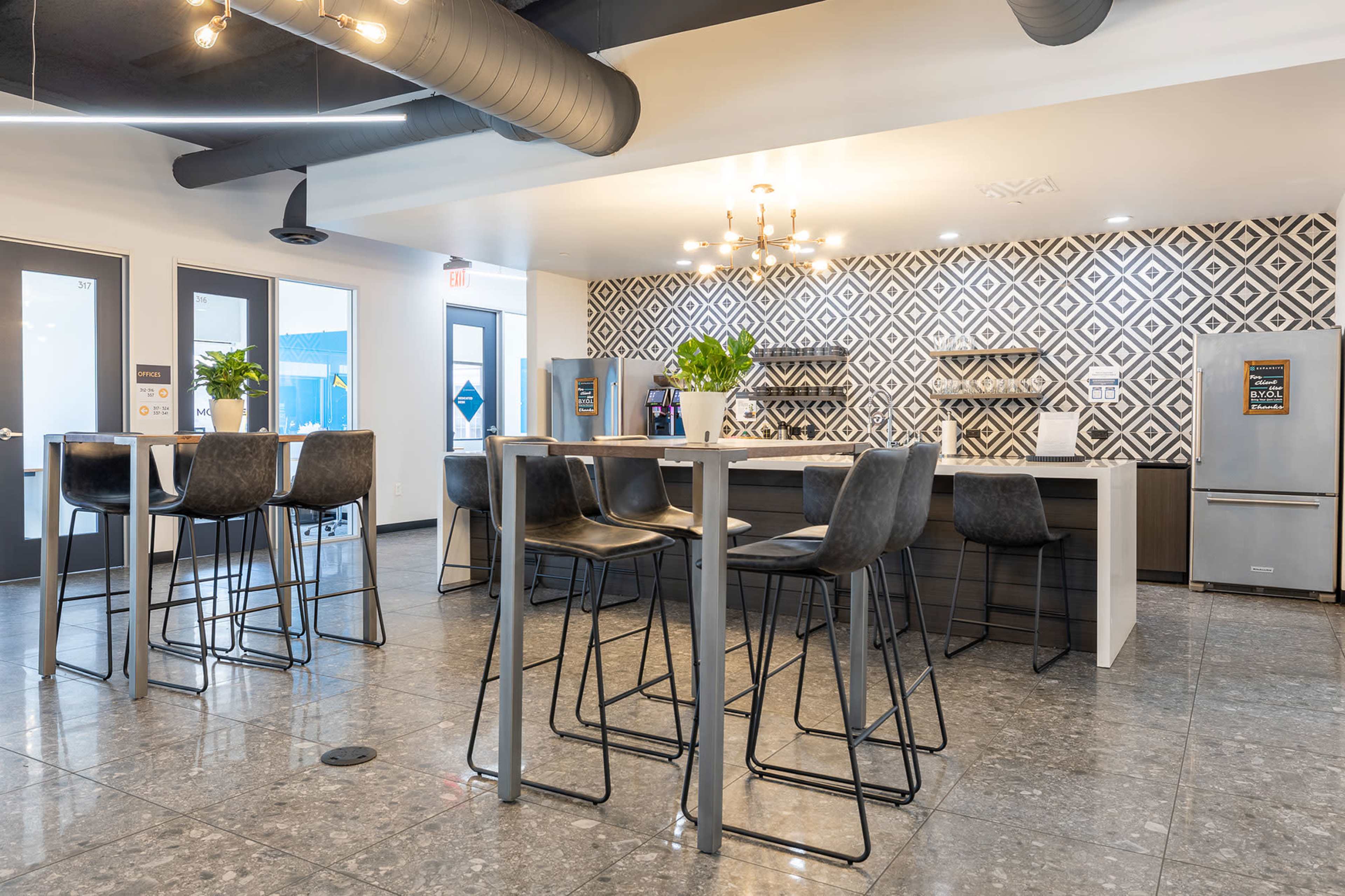 The image shows a modern kitchen and dining area with high tables and chairs, featuring a patterned backsplash and stainless steel appliances.
