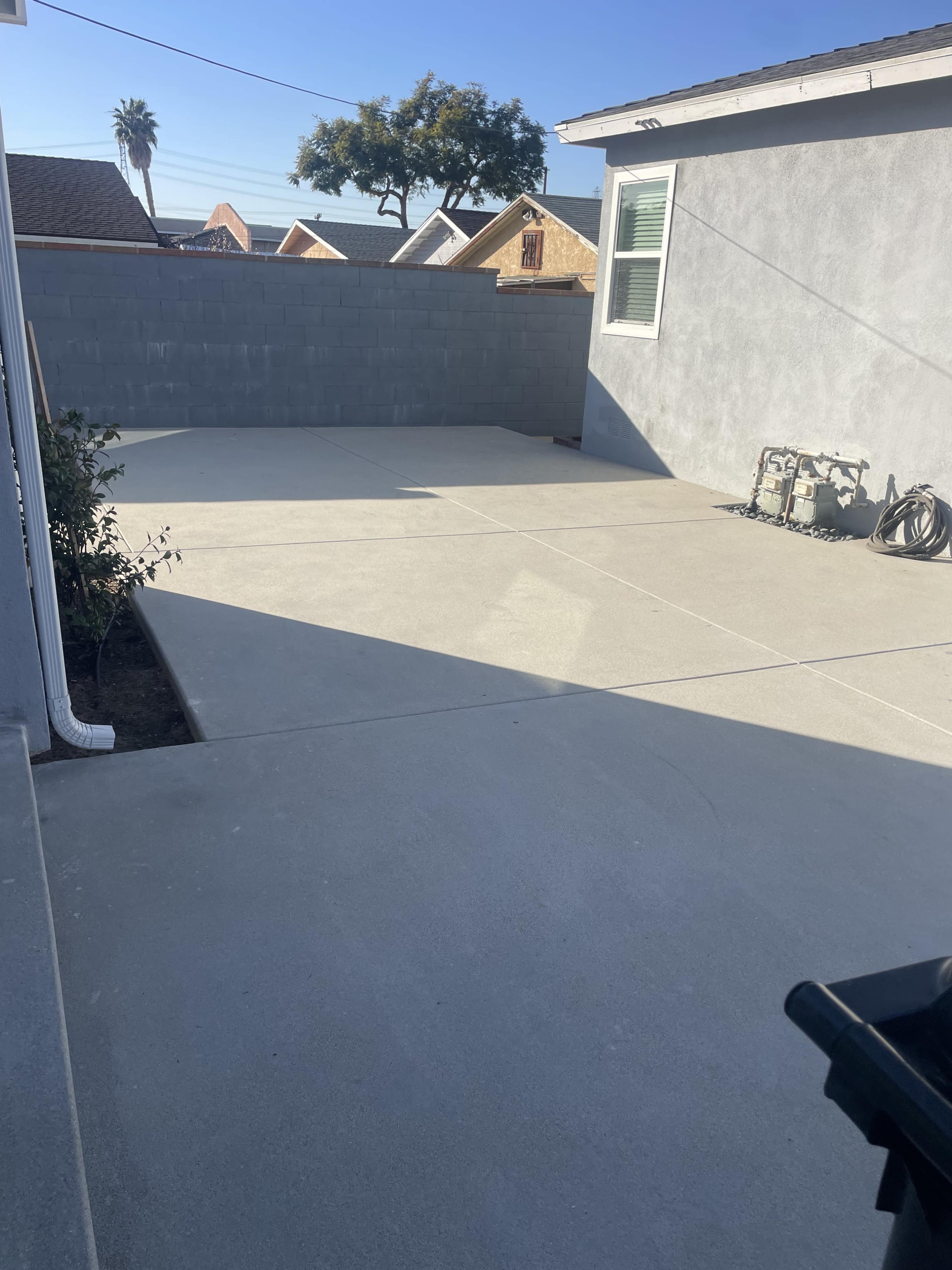 The image shows a concrete outdoor area next to a house, bordered by a gray wall and featuring a few plants and utility pipes.