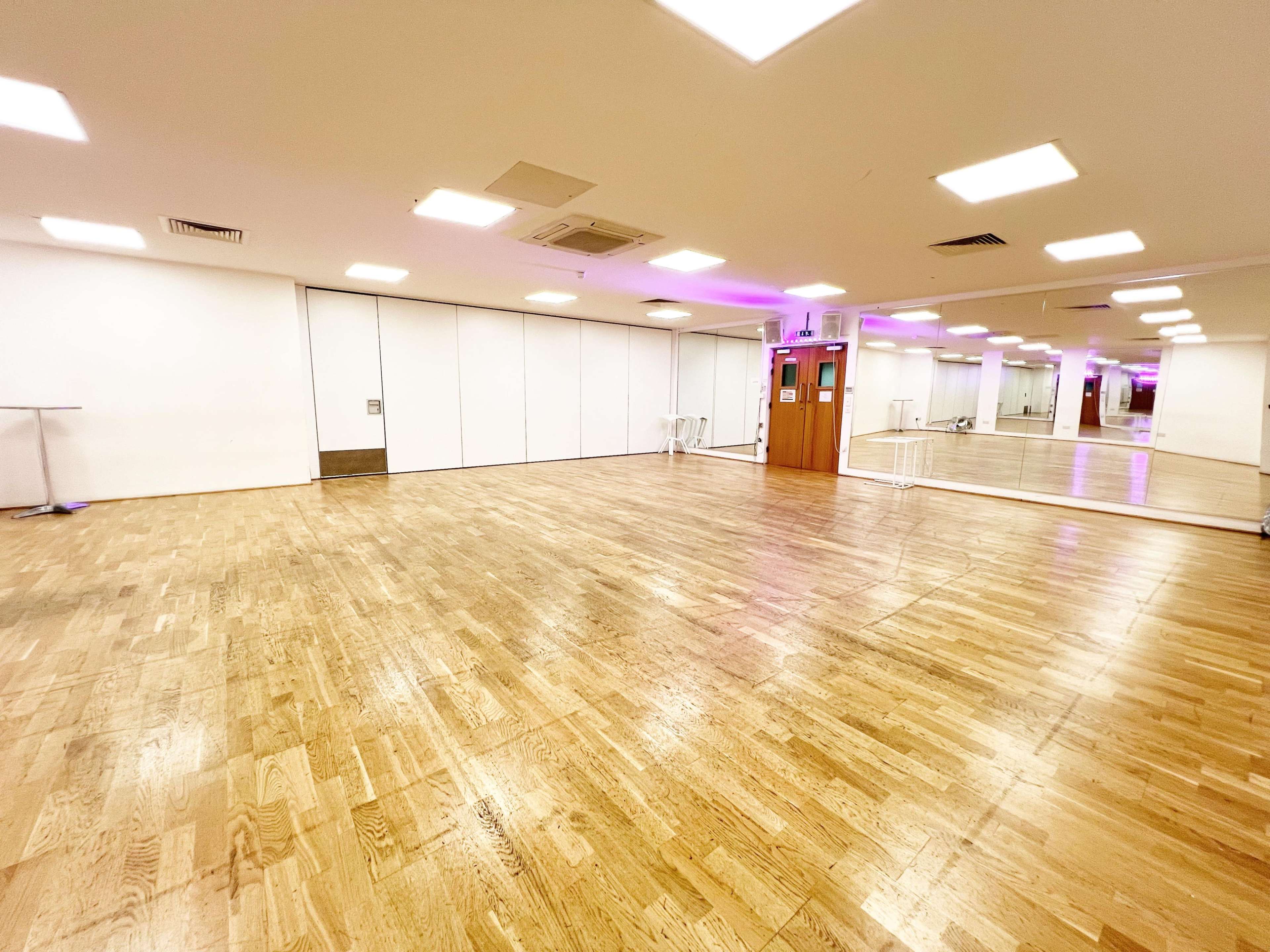 The image shows a spacious empty dance studio with wooden flooring and mirrors on one wall.