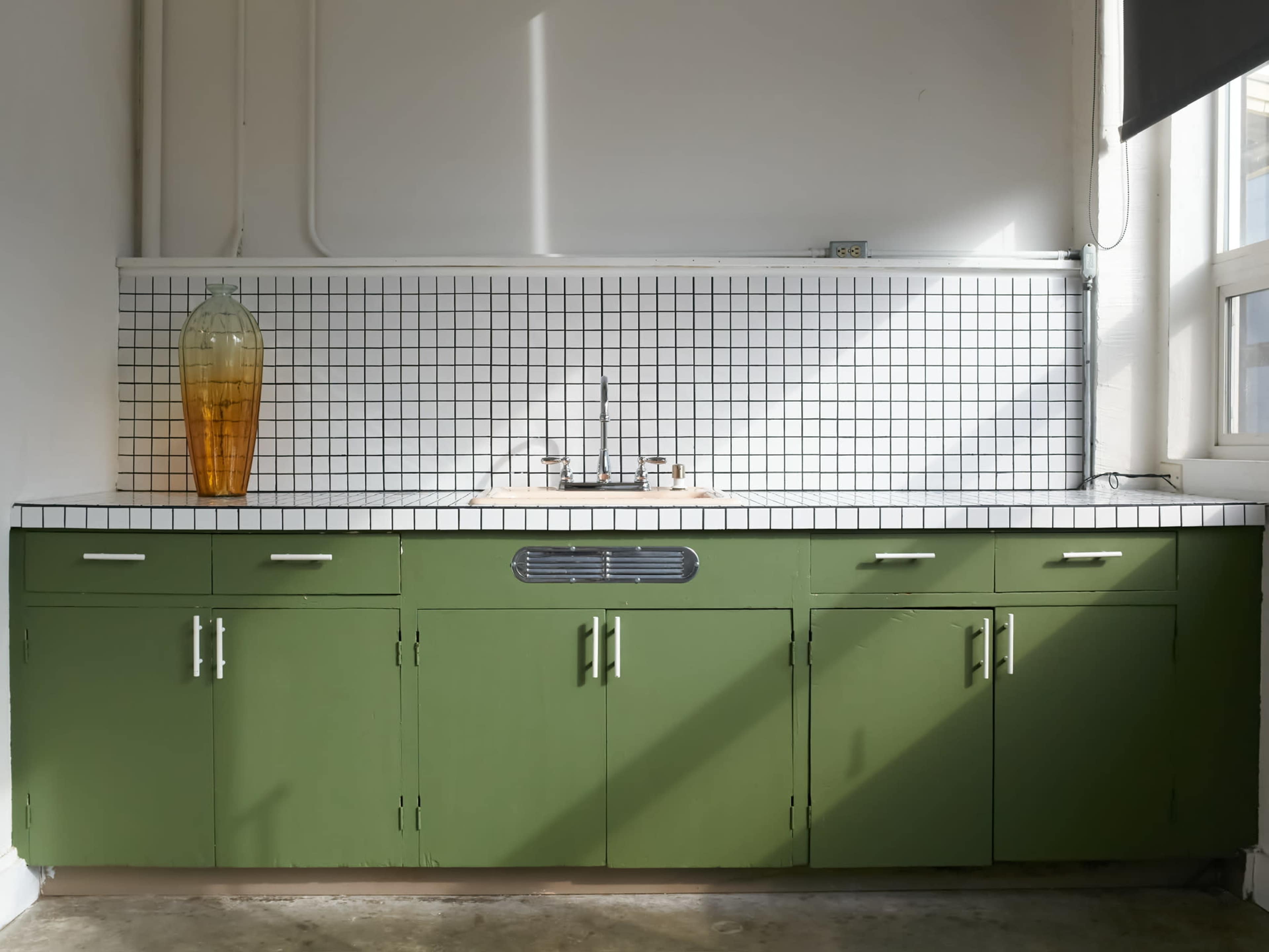 The image shows a kitchen featuring green cabinetry with a white tiled backsplash and a large amber vase on the counter.