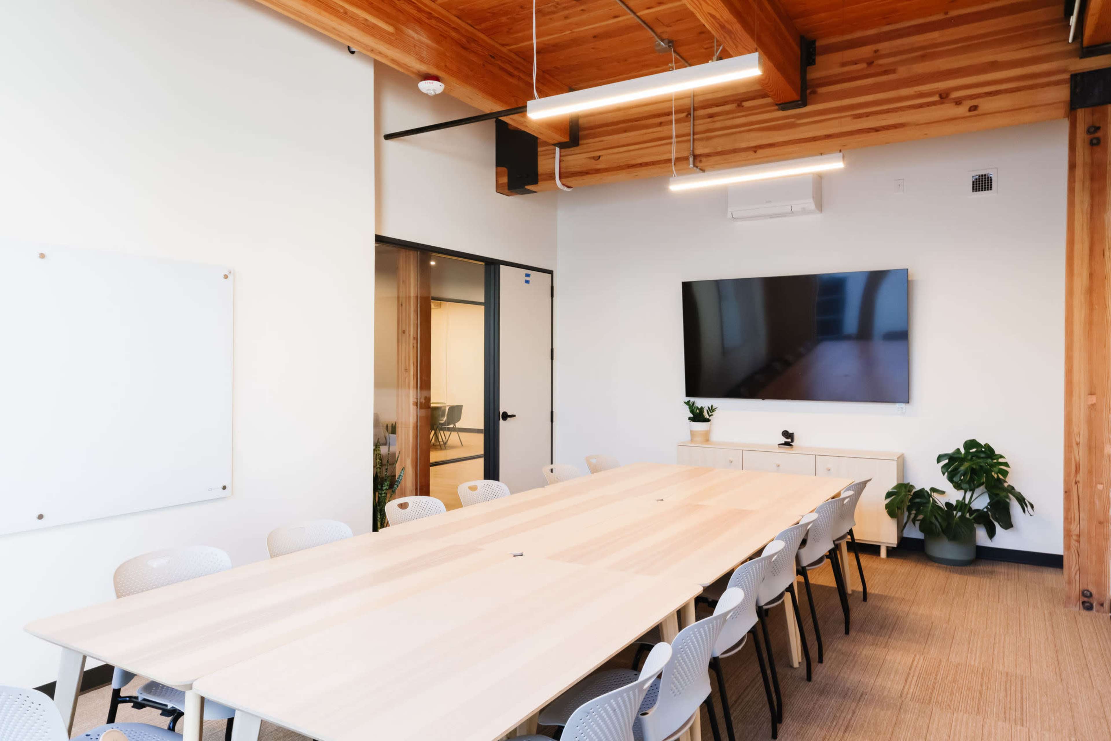 A modern conference room with a long wooden table, several light gray chairs, a large wall-mounted television, and potted plants.