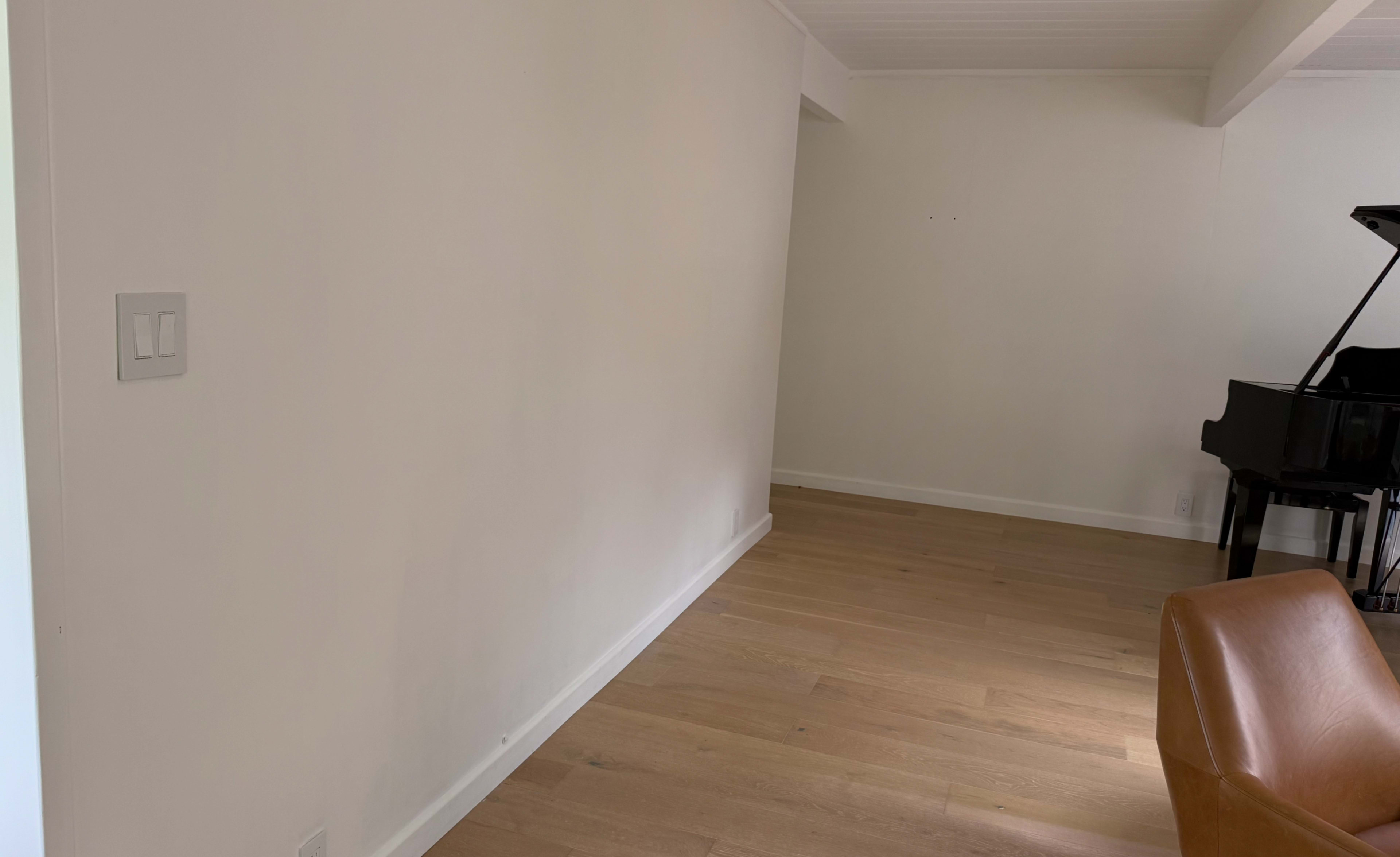 The image shows a minimalist room with a light wood floor, an empty white wall, and a black piano in the corner.