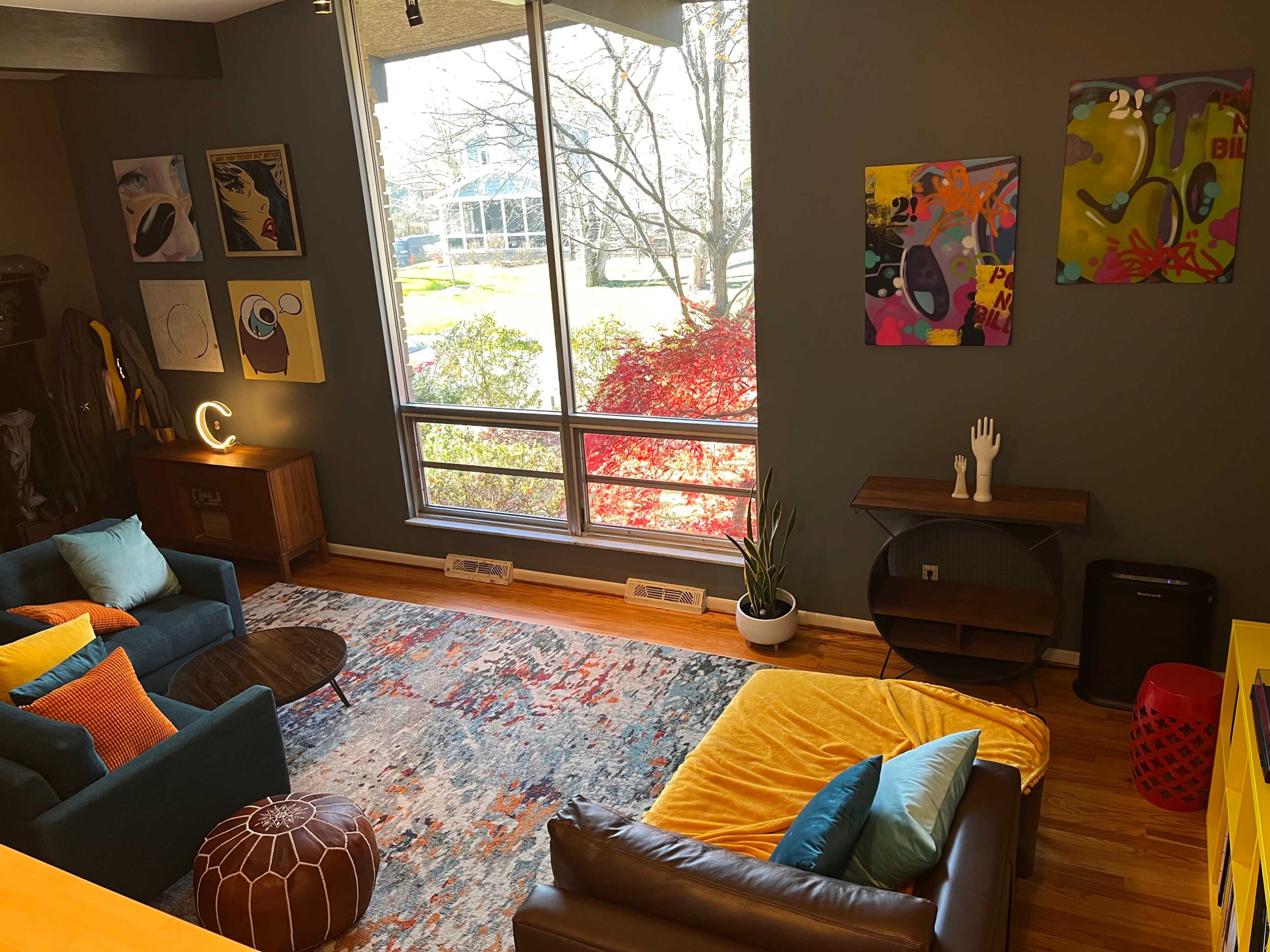 A cozy living room with two sofas, a colorful rug, and large windows letting in natural light, featuring vibrant artwork on the walls.