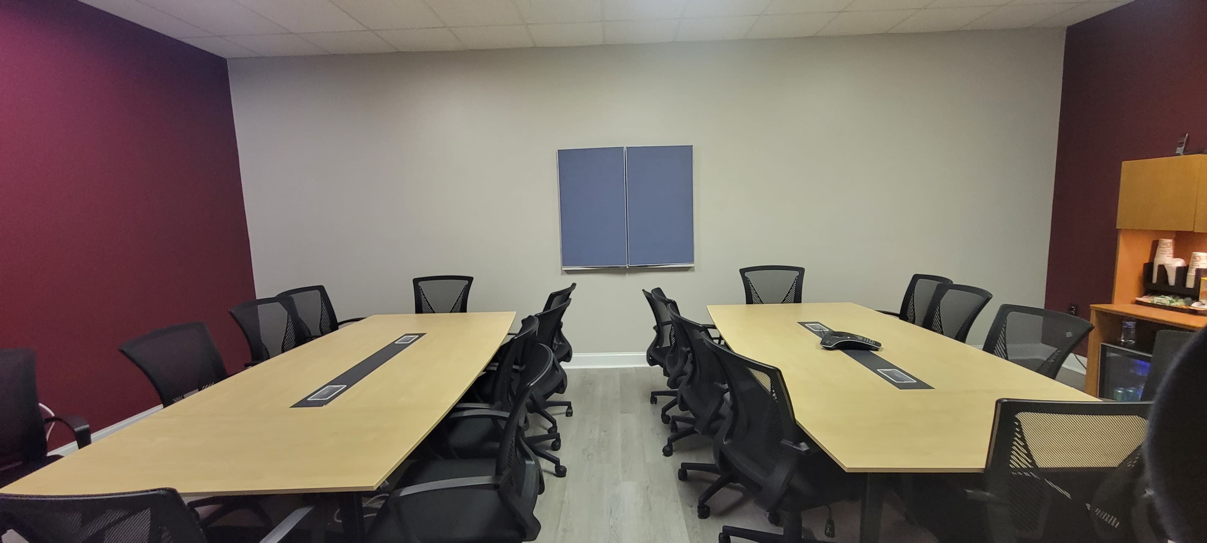A conference room features two long tables arranged in a U-shape with black chairs and a blank wall art piece.