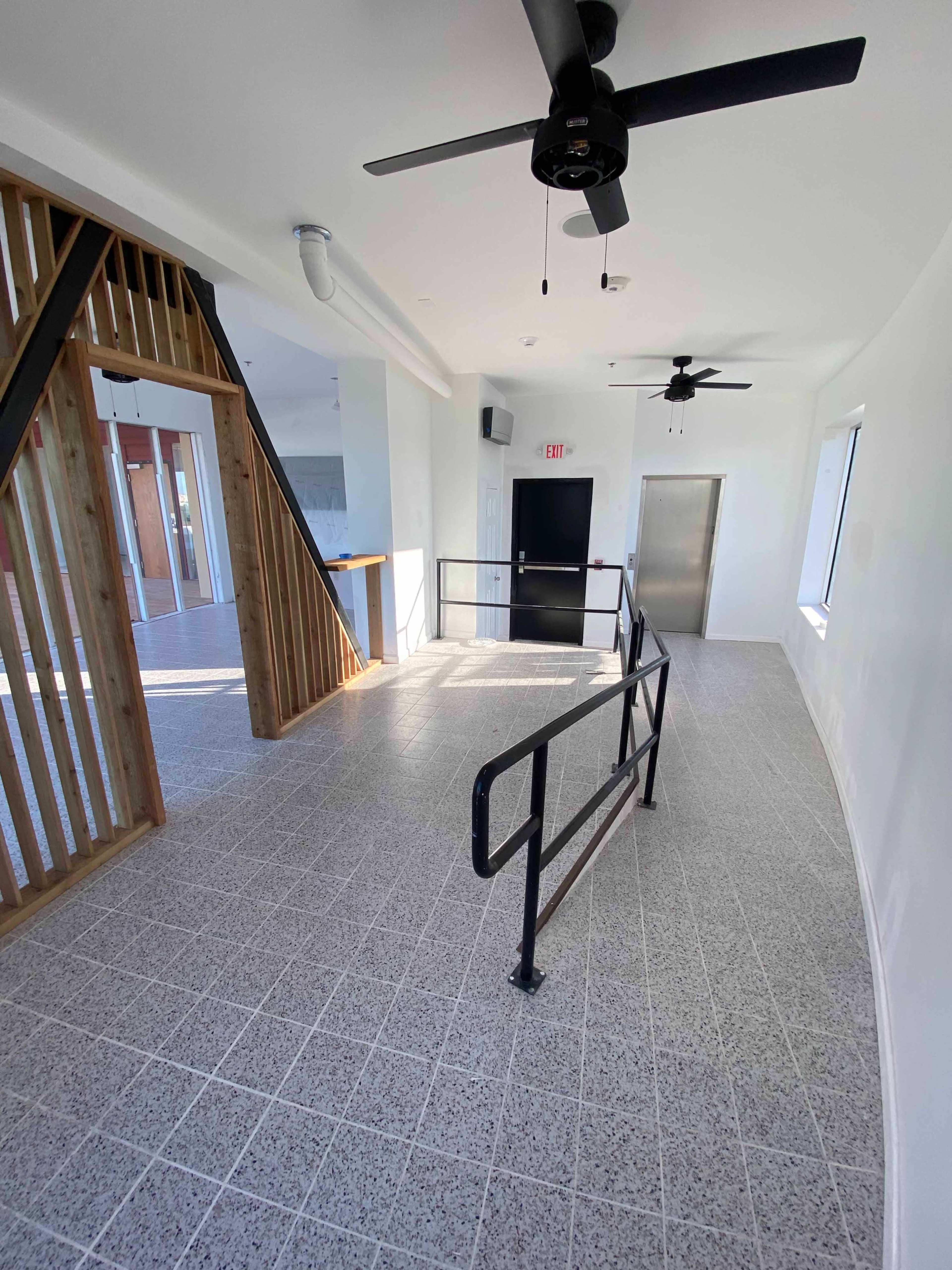 A well-lit corridor with a tiled floor, featuring black railings and ceiling fans, leading to a door and an elevator.