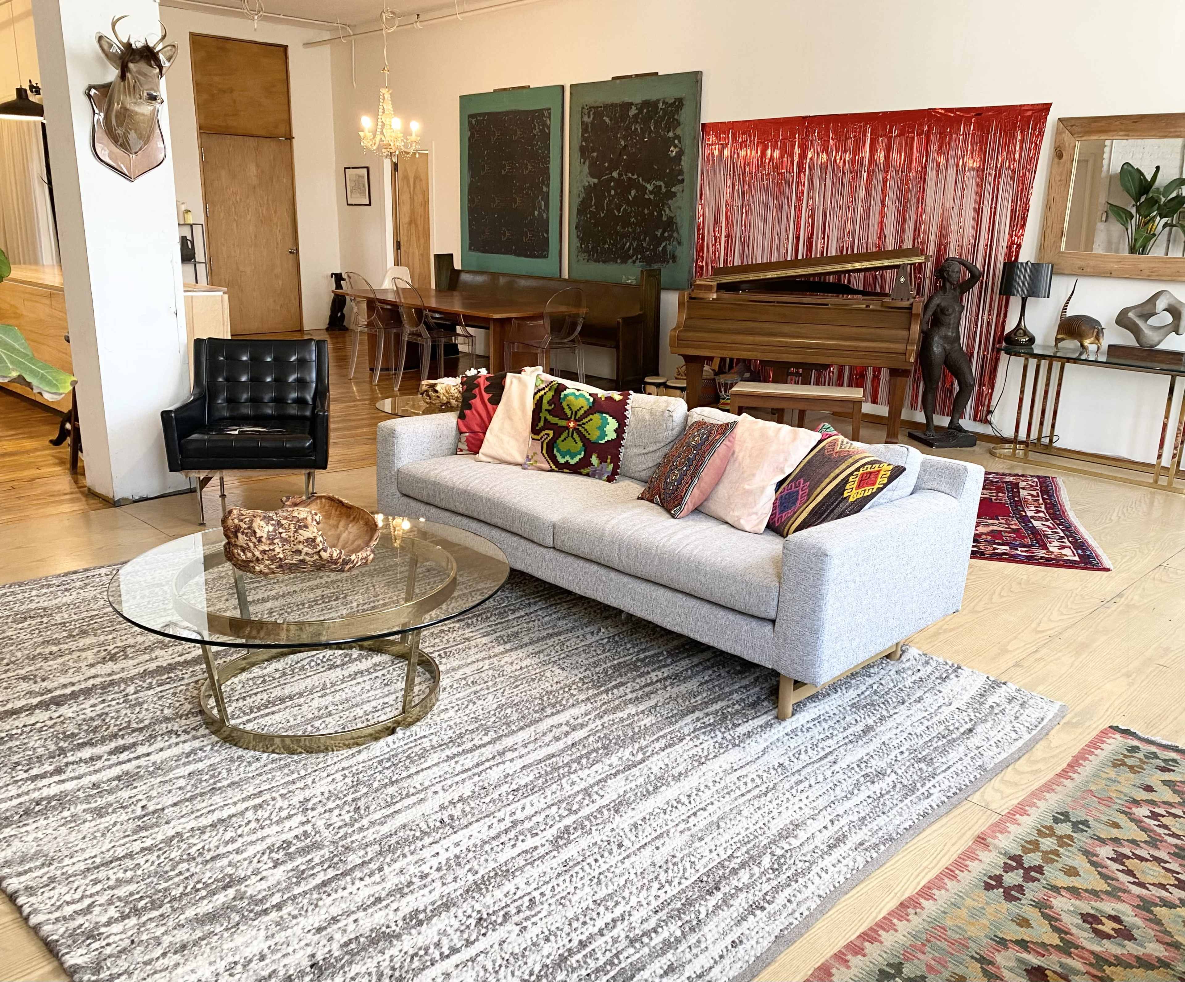 A modern living space featuring a gray sofa with colorful cushions, a round glass coffee table, and a piano, surrounded by a mix of decorative rugs and artwork.