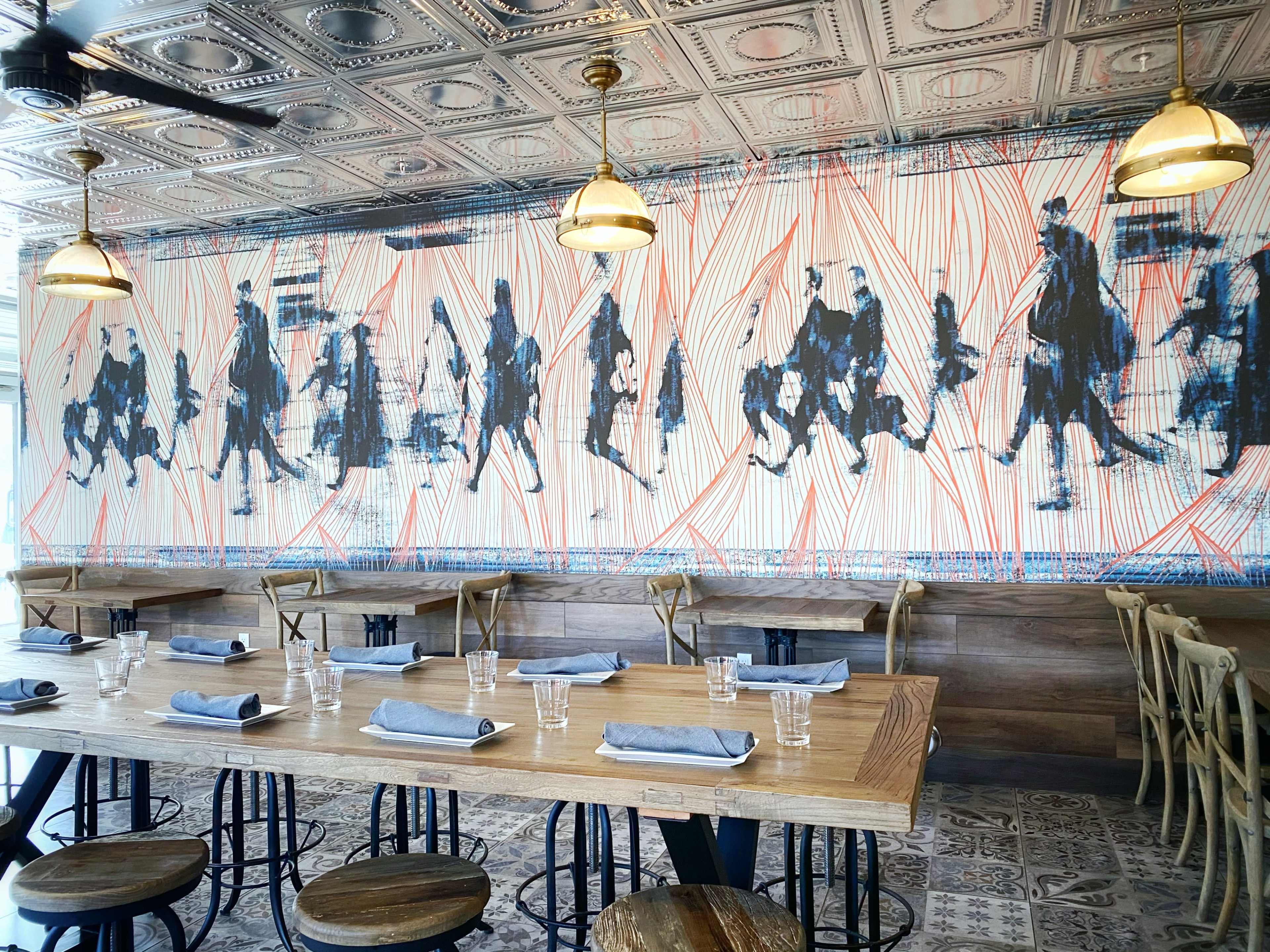 A restaurant interior with a wooden table set for dining, featuring a mural of soldiers on a backdrop of red lines.