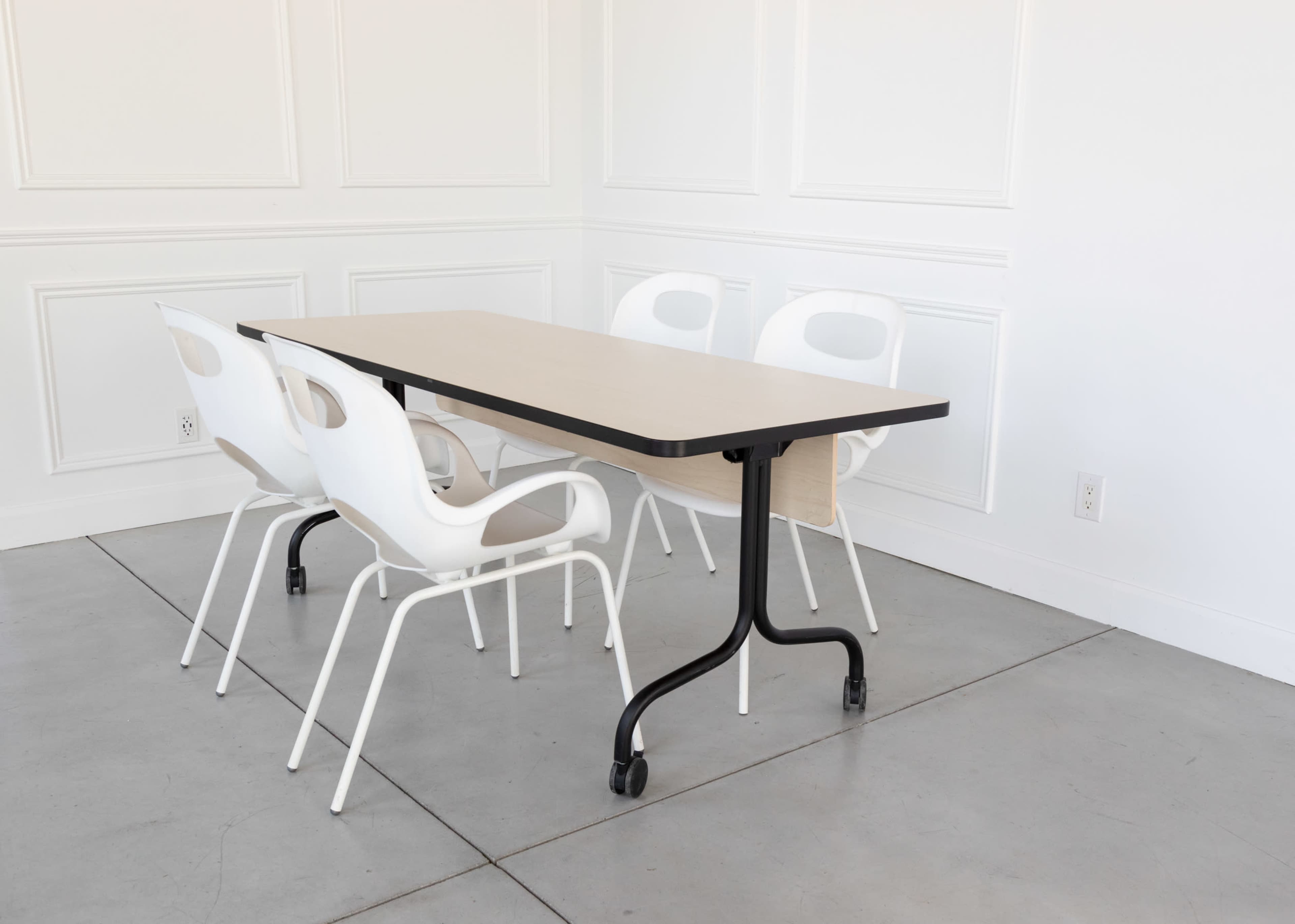 A rectangular table with a light tabletop and black legs is accompanied by four white, wheeled chairs on a concrete floor in a minimalist room.