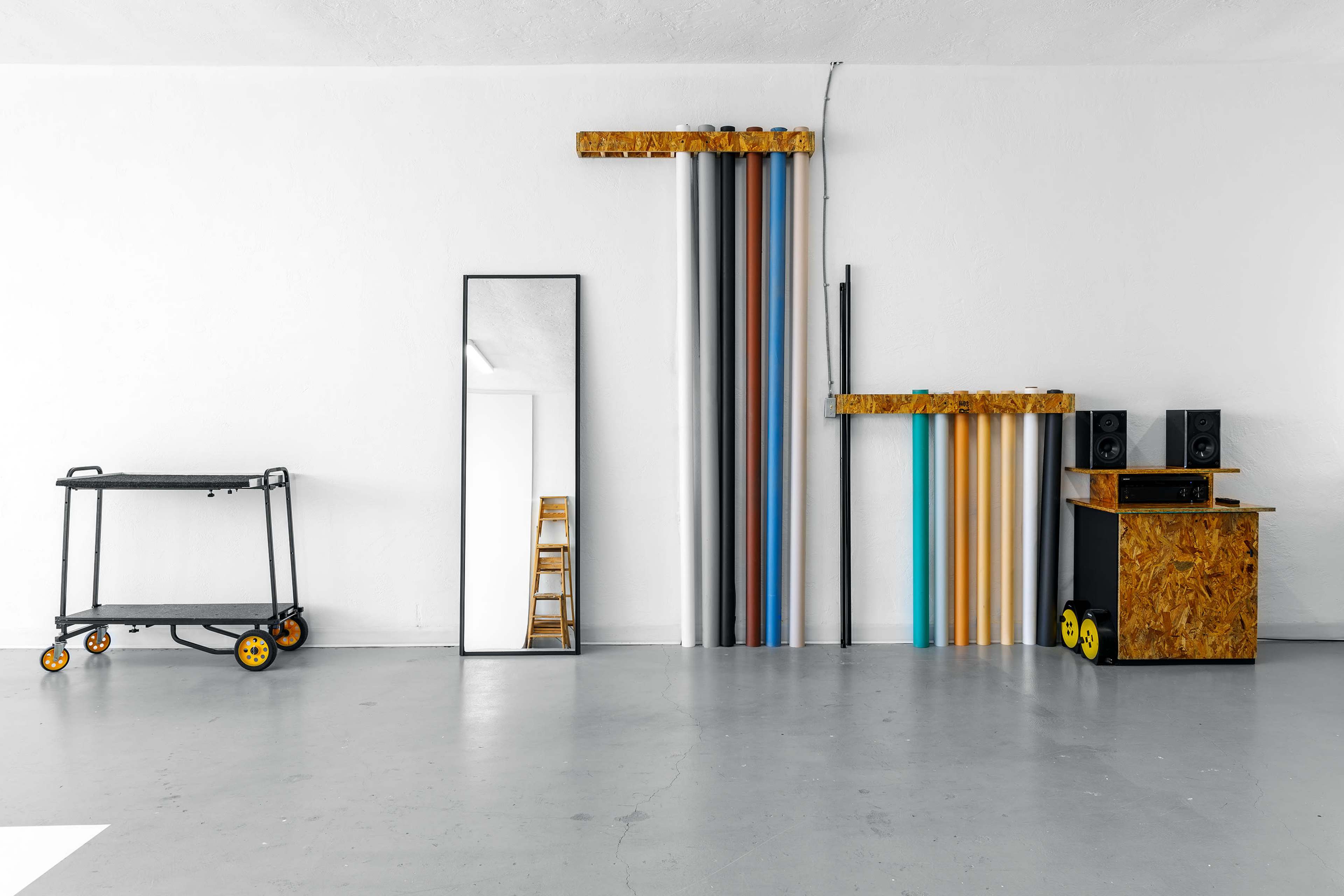 A modern studio space featuring a mirrored cabinet, various rolls of colored material on the wall, a wooden cart on wheels, and a small wooden desk with speakers.