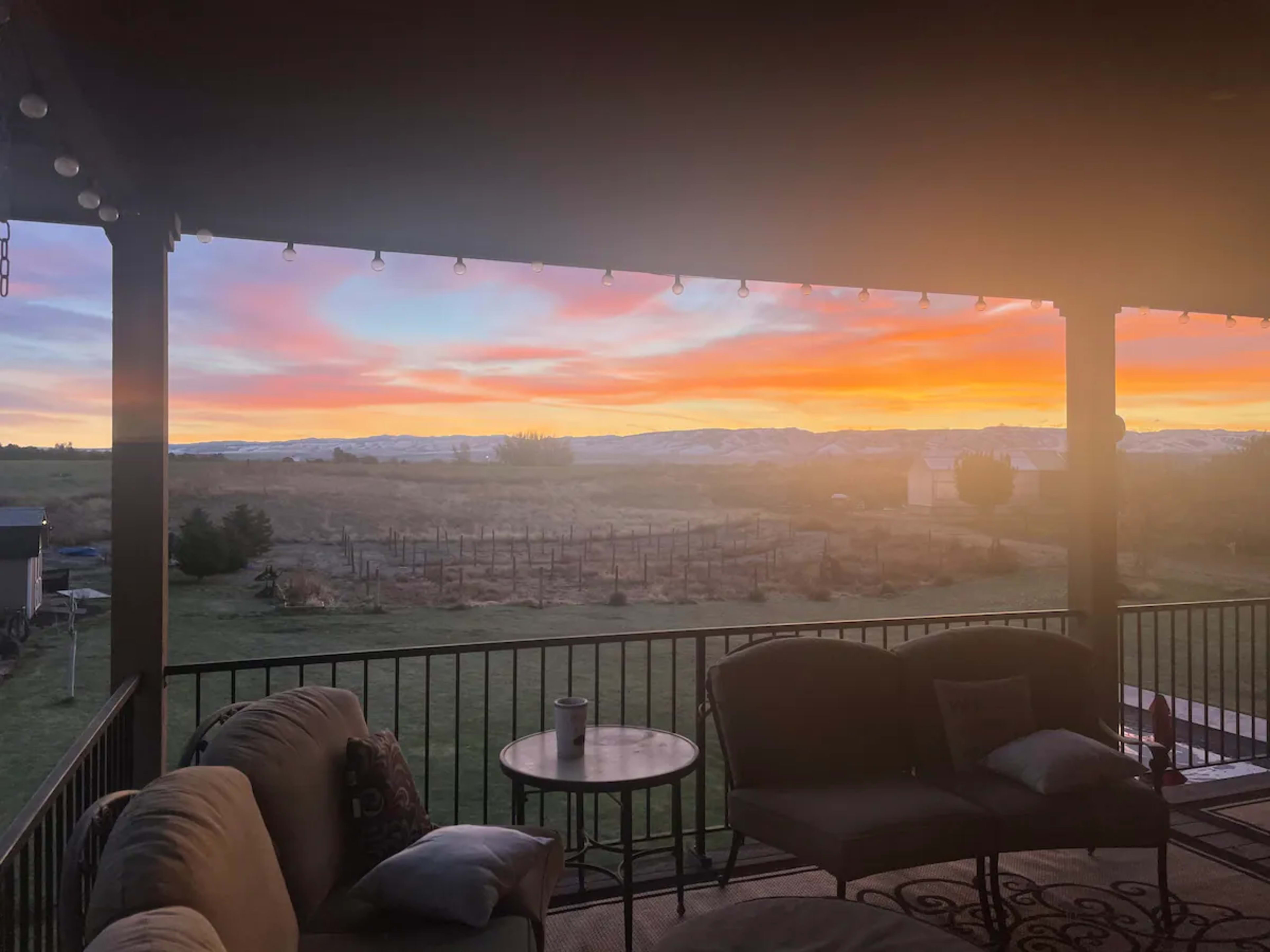 The image shows a patio area with a couch and table overlooking a vibrant sunset over distant mountains and a vineyard.