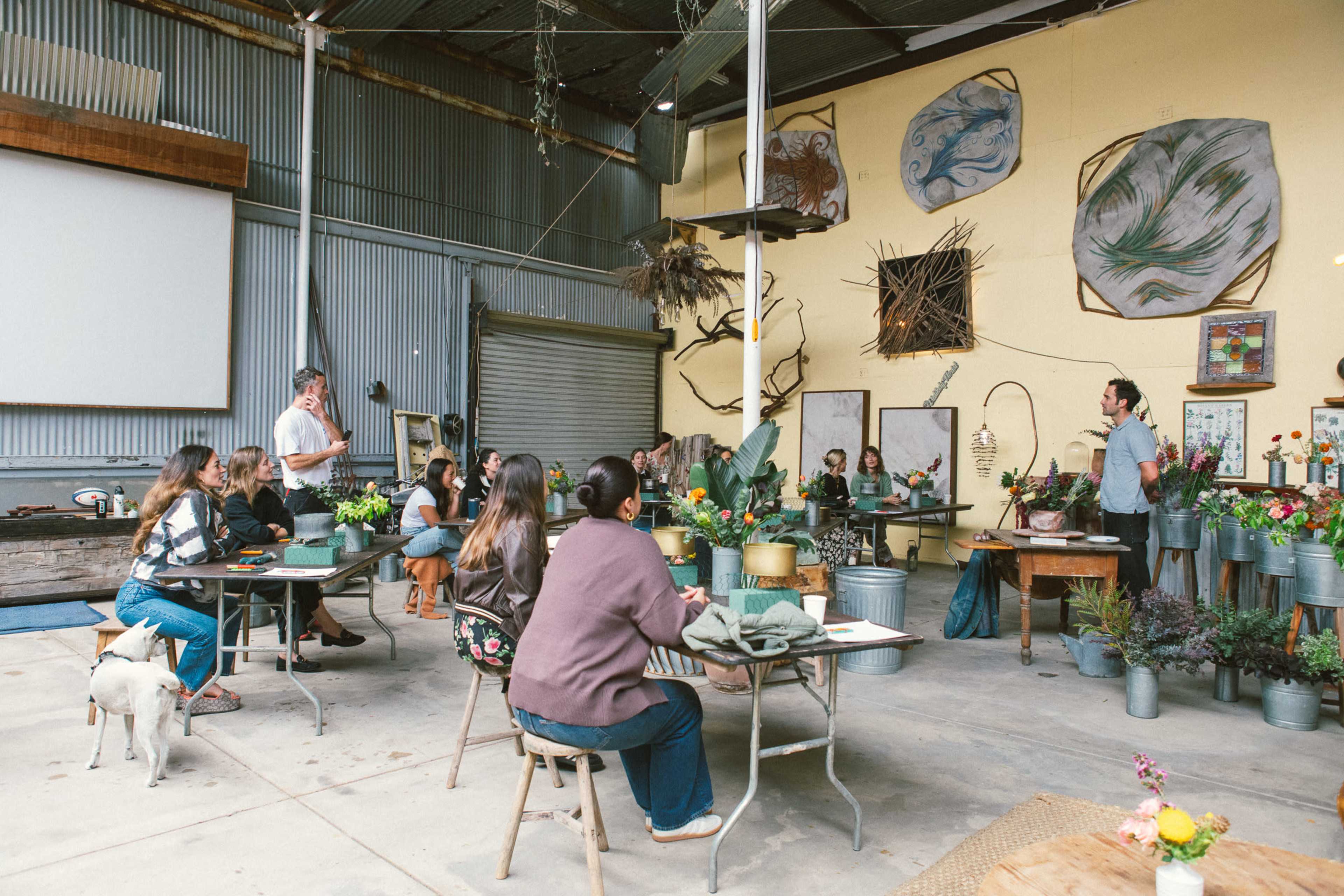 A group of people sits at tables in a workshop space filled with plants and artistic decorations while someone speaks at the front.